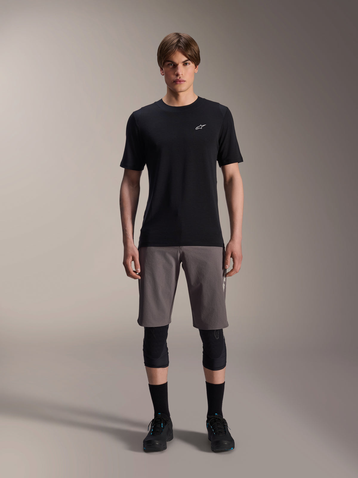 Alpinestars A-Dura Dri Wool Short Sleeve Jersey, Black, worn by a model with grey mountain bike shorts, black knee guards, and black cycling shoes, featuring a minimalist design with a small white Astars logo on the chest, functional performance MTB apparel for trail riding.