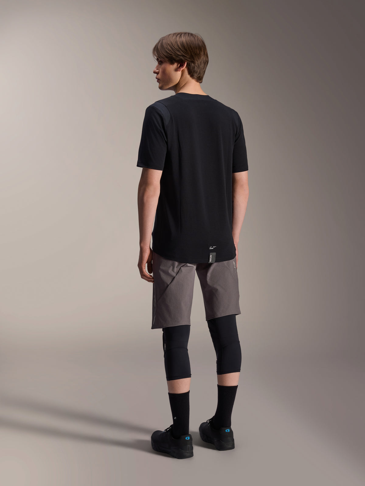 Alpinestars A-Dura Dri Wool Short Sleeve Jersey, Black, back view on model, worn with grey mountain biking shorts and black knee guards, featuring a dropped tail hem and subtle logo, technical MTB apparel