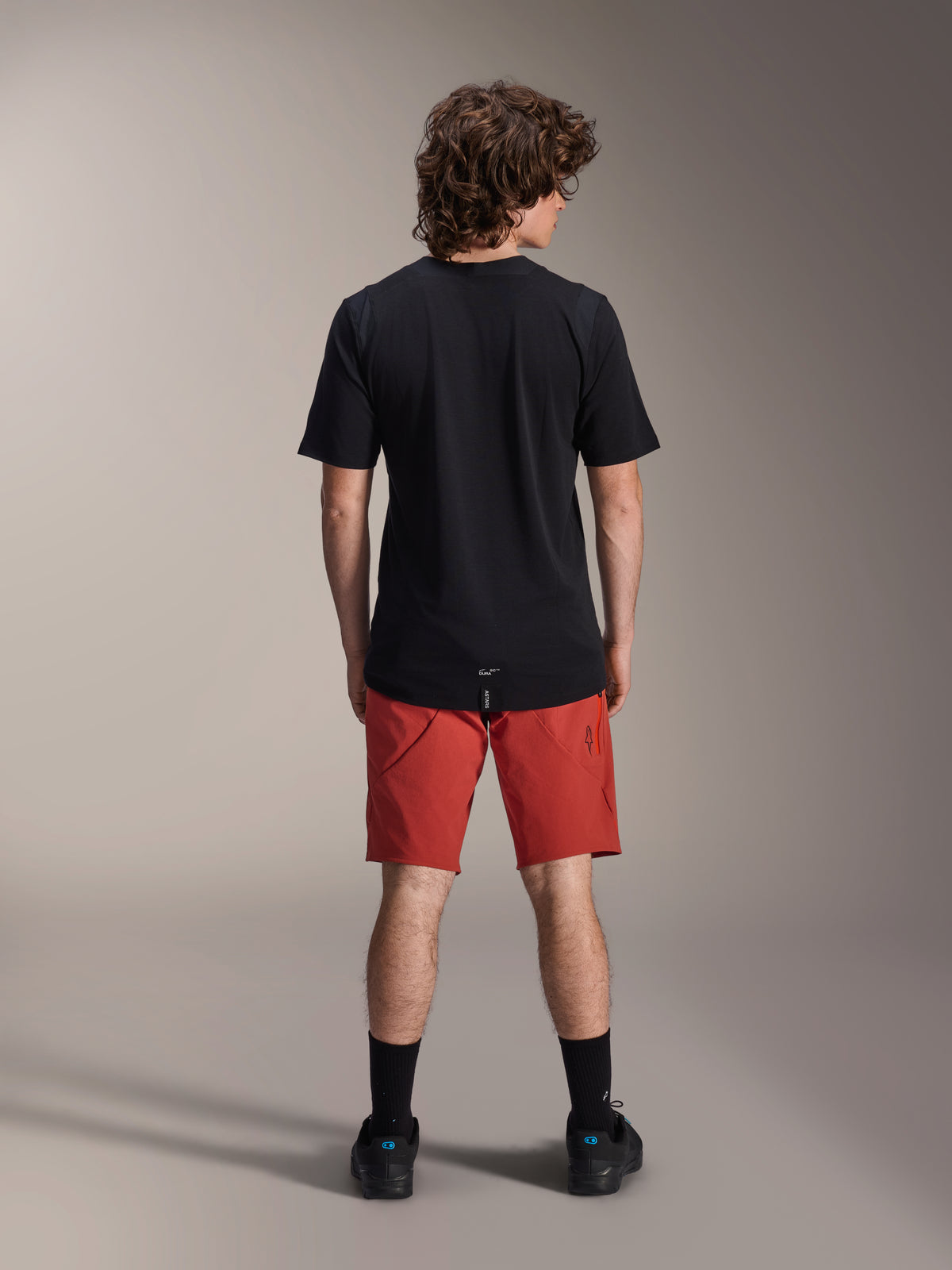 Modèle masculin portant le short Alpinestars A-Dura Elite orange foncé et le maillot à manches courtes A-Dura Dri Wool noir, vu de dos, avec chaussettes noires et chaussures de cyclisme, tenue de VTT professionnelle