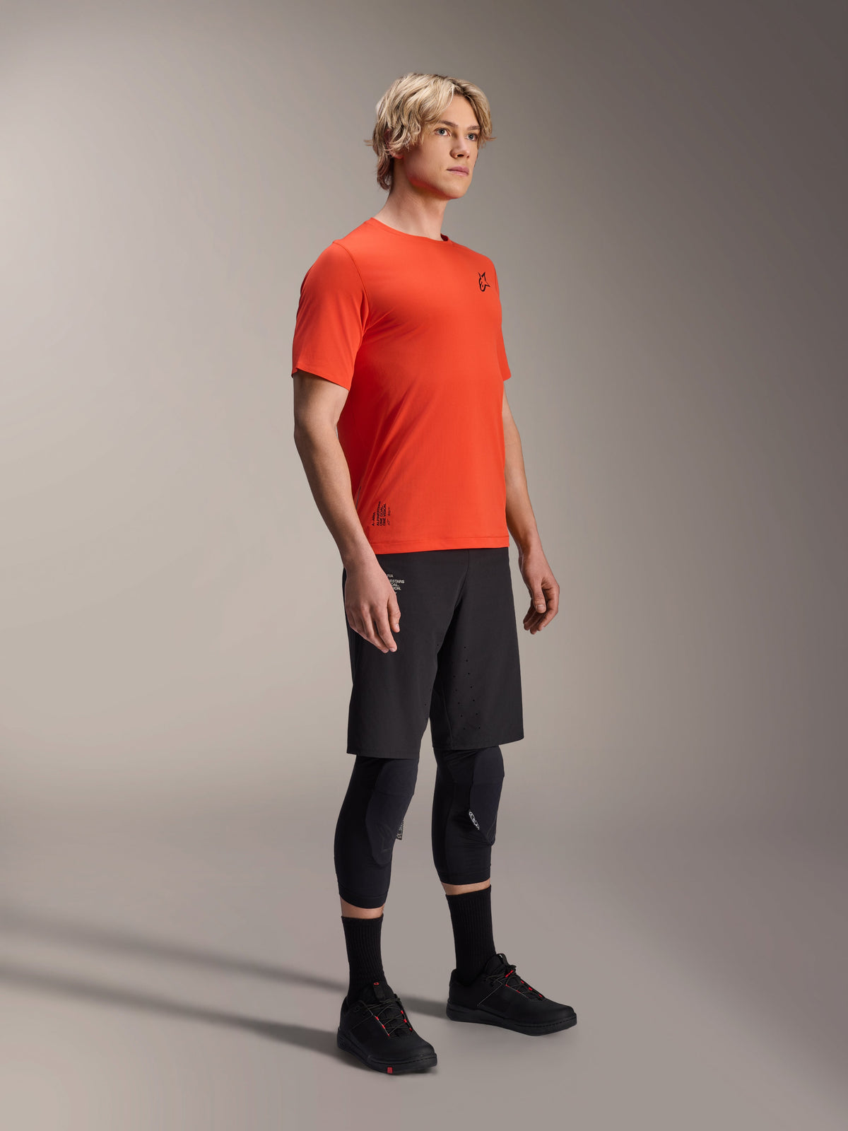 Alpinestars A-Aria Astar Short Sleeve Jersey in Spicy Orange, worn by a model with black MTB shorts and knee protection, lightweight breathable mountain bike apparel, vibrant orange with minimal black branding