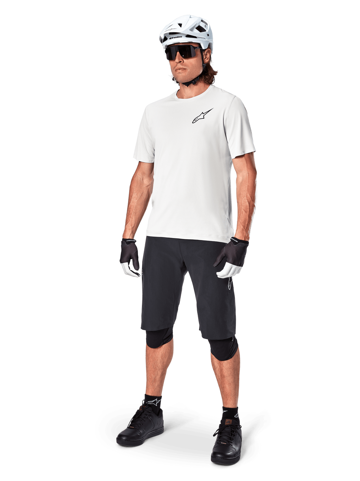 Cycliste VTT portant un maillot à manches courtes Alpinestars A-Aria Astar en White Dove, associé à un short de VTT noir, un casque blanc et des genouillères noires, avec un petit logo noir sur la poitrine, équipement de cyclisme coordonné pour la performance sur sentier