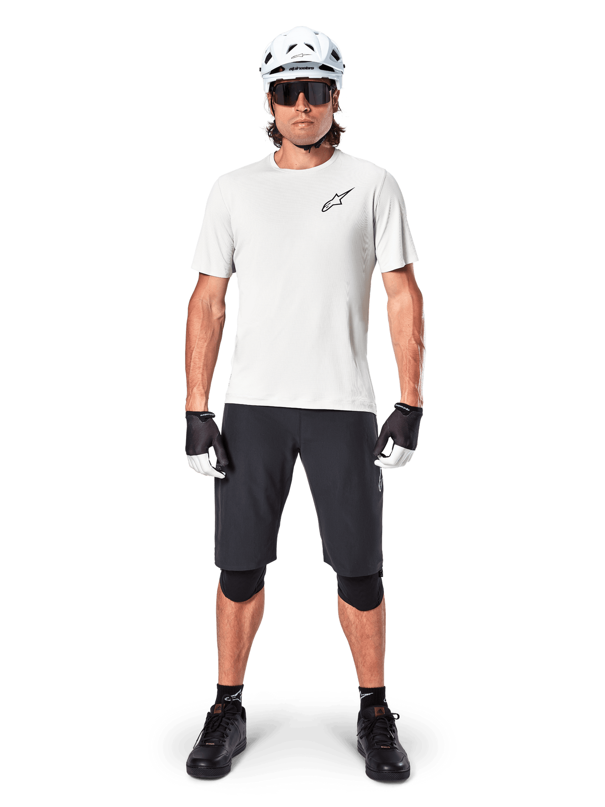 Cycliste portant le maillot à manches courtes Alpinestars A-Aria Astar en coloris White Dove, avec un logo Astars noir sur la poitrine, assorti d'un short de VTT noir, d'un casque blanc, de lunettes de soleil, de genouillères noires et de gants, tenue de cyclisme tout-terrain complète.