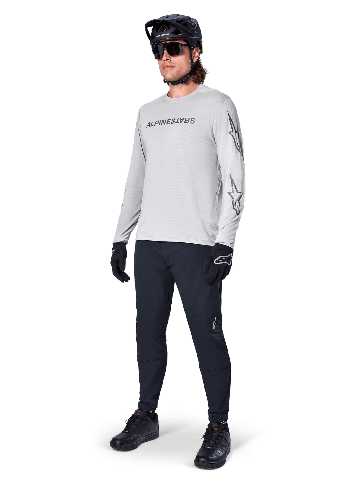 Maillot à manches longues Alpinestars A-Dura Switch, gris clair, porté par un cycliste avec un pantalon de VTT noir et un casque, arborant un logo lettrage noir sur la poitrine et des graphismes d'étoiles sur les manches, équipement de cyclisme tout-terrain haute performance