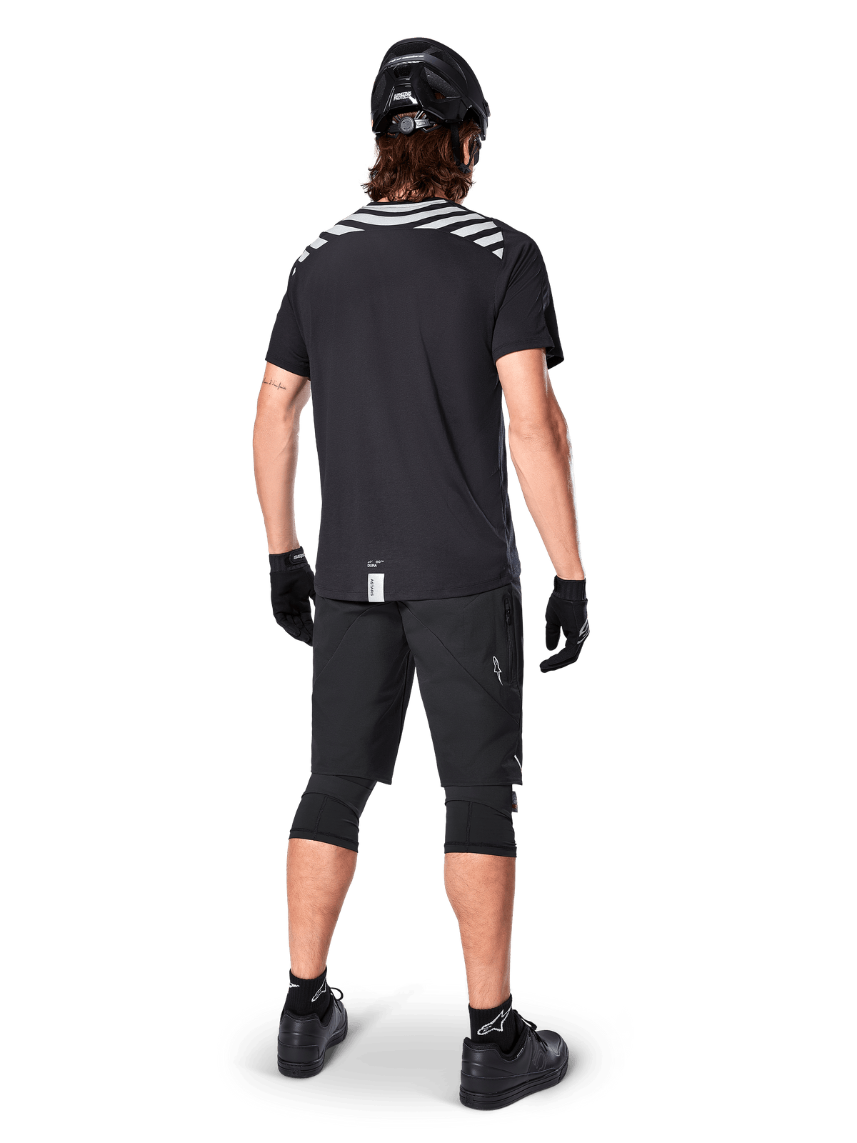 Alpinestars A-Dura Dri Oscar Short Sleeve Jersey, Black Reflective, rear view of a rider wearing the jersey with black mountain bike shorts, helmet, and knee pads, featuring reflective shoulder graphics and a drop tail design for MTB riding.