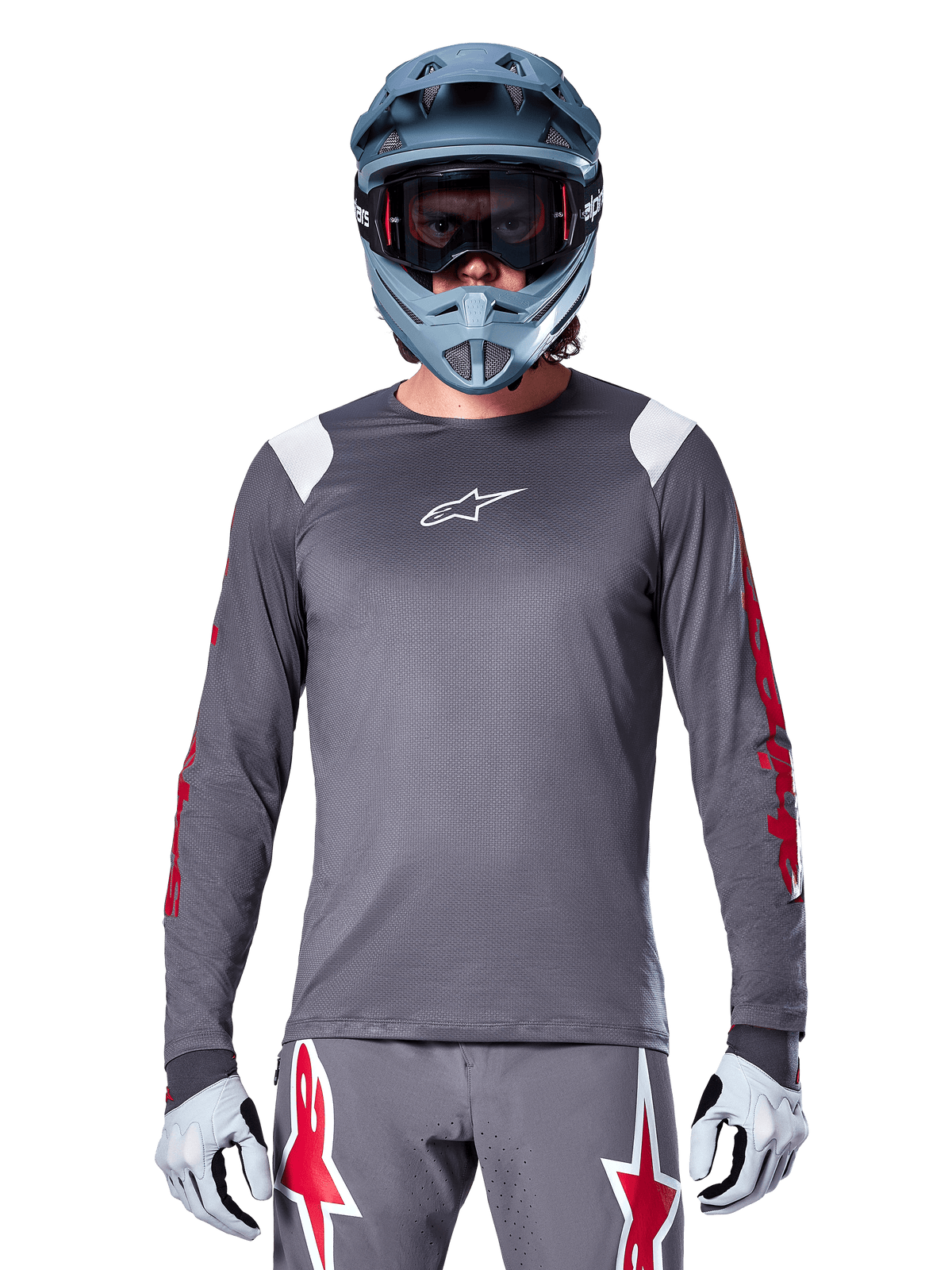 Cycliste VTT portant le maillot à manches longues Alpinestars A-Supra Astar, gris foncé avec graphismes rouges sur les manches et accents blancs aux épaules, pantalon gris assorti avec logos étoiles rouges, casque intégral bleu et masque, équipement de cyclisme tout-terrain coordonné