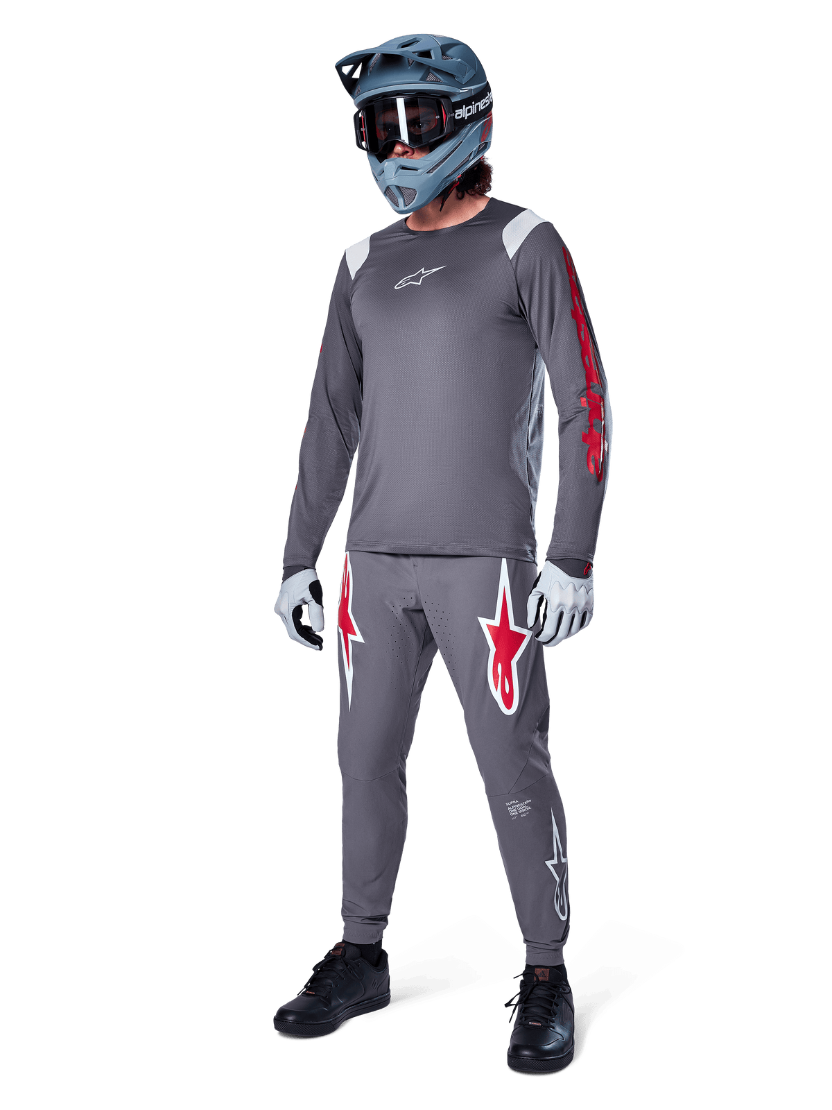Maillot à manches longues Alpinestars A-Supra Astar gris foncé, avec marquage rouge sur les manches et logo blanc sur la poitrine, porté par un cycliste avec un pantalon gris assorti, un casque intégral bleu-gris et des gants de performance, tenue de VTT coordonnée pour la pratique de la gravité.