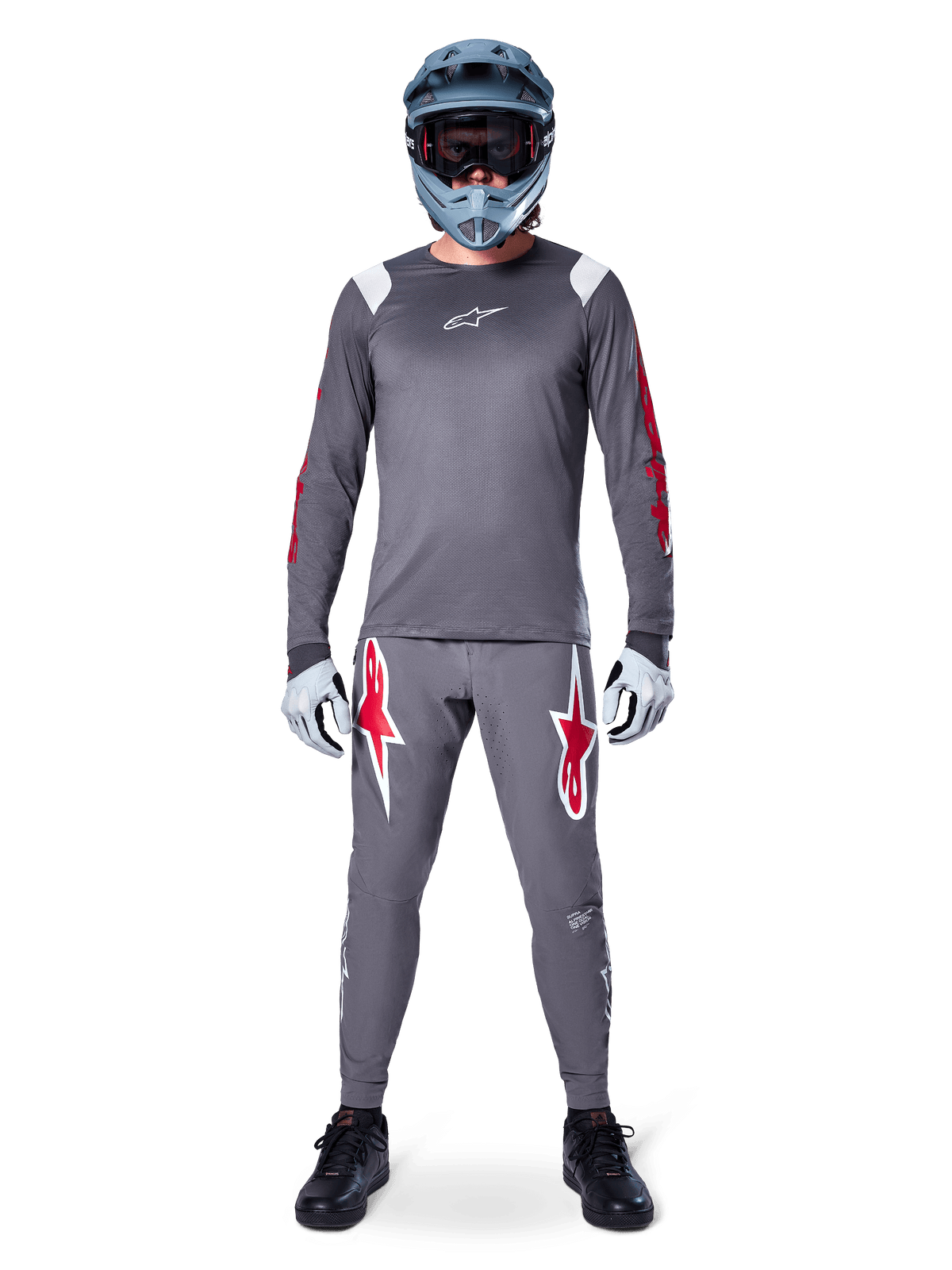 Pilote de VTT portant le maillot à manches longues Alpinestars A-Supra Astar, gris foncé avec des accents rouges et un logo blanc sur la poitrine, pantalon gris assorti avec de grands graphismes d'étoiles rouges, casque intégral bleu avec masque, gants gris clair et chaussures noires, ensemble d'équipement de VTT professionnel