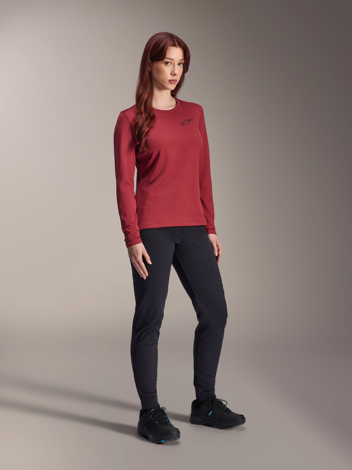 Maillot thermique Alpinestars Stella A-Dura, maillot de VTT à manches longues pour femme, bordeaux, porté avec un pantalon de sport noir et des chaussures noires, col rond avec un petit logo noir sur la poitrine, design à coupe régulière et fonctionnelle pour le cyclisme tout-terrain