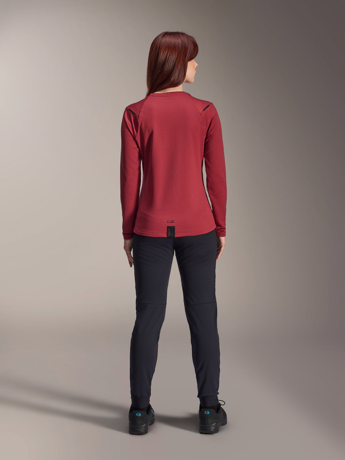 Femme portant le maillot Alpinestars Stella A-Dura Thermal, Bordeaux, vue arrière, maillot de cyclisme thermique à manches longues avec pan arrière allongé, associé à un pantalon noir et des chaussures de vélo noires, tenue de VTT