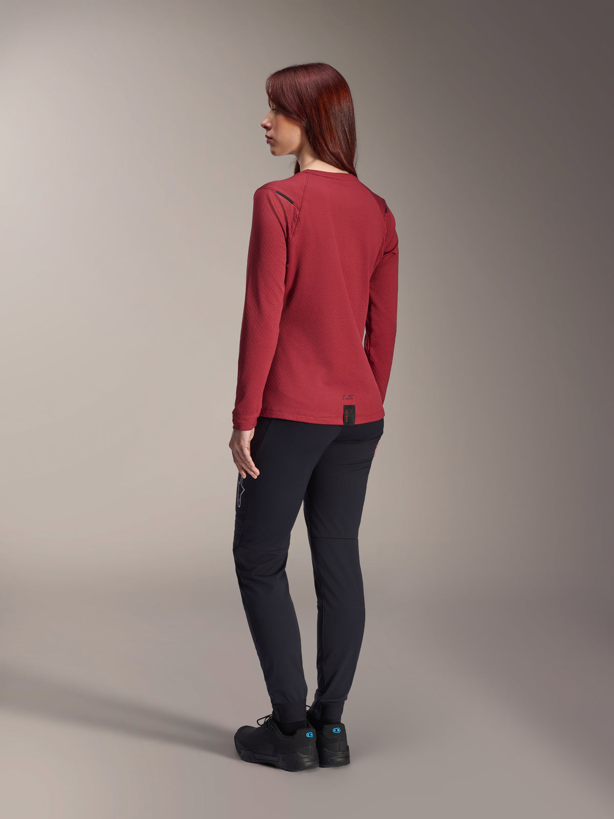 Alpinestars Stella A-Dura Thermal Jersey, maillot de VTT à manches longues pour femme, bordeaux, vue de dos mettant en avant le pan arrière allongé et la texture du tissu thermique, porté avec un pantalon de VTT noir et des chaussures noires