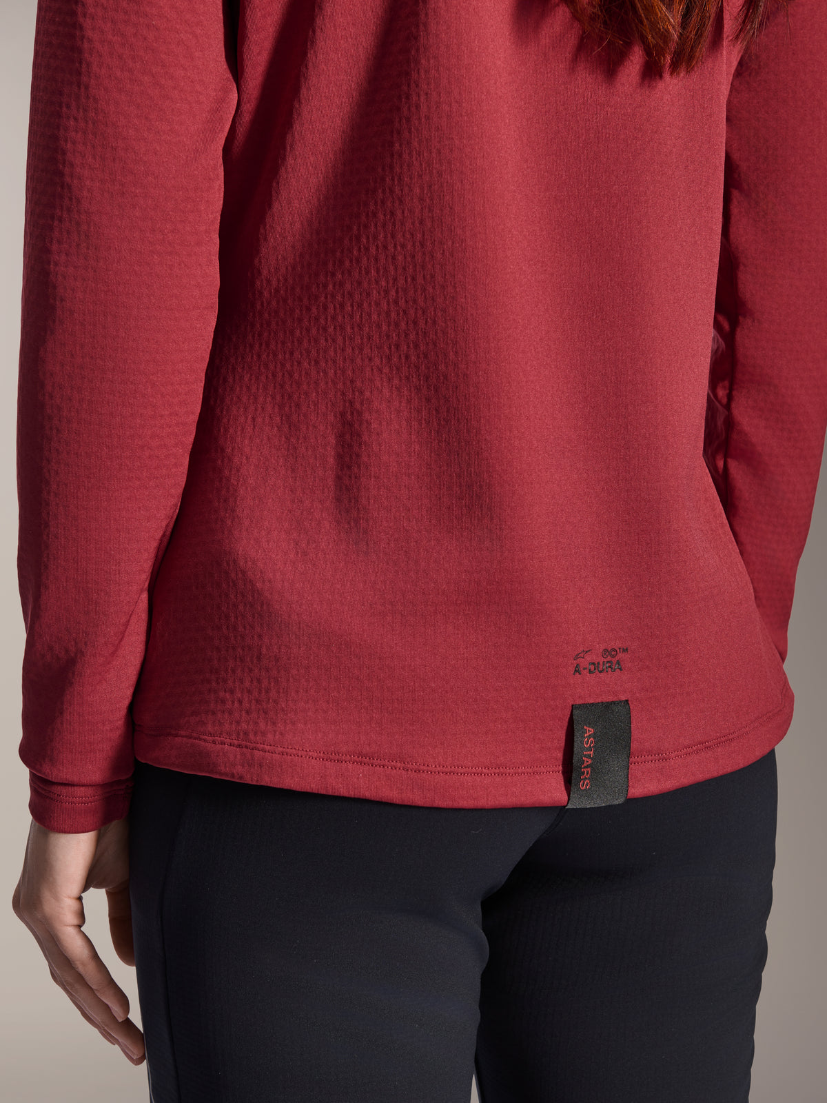 Maillot thermique femme Alpinestars Stella A-Dura, bordeaux, vue rapprochée de l'ourlet arrière présentant le marquage A-Dura et l'étiquette tissée ASTARS, tissu polaire isolant texturé, vêtement de VTT pour femme