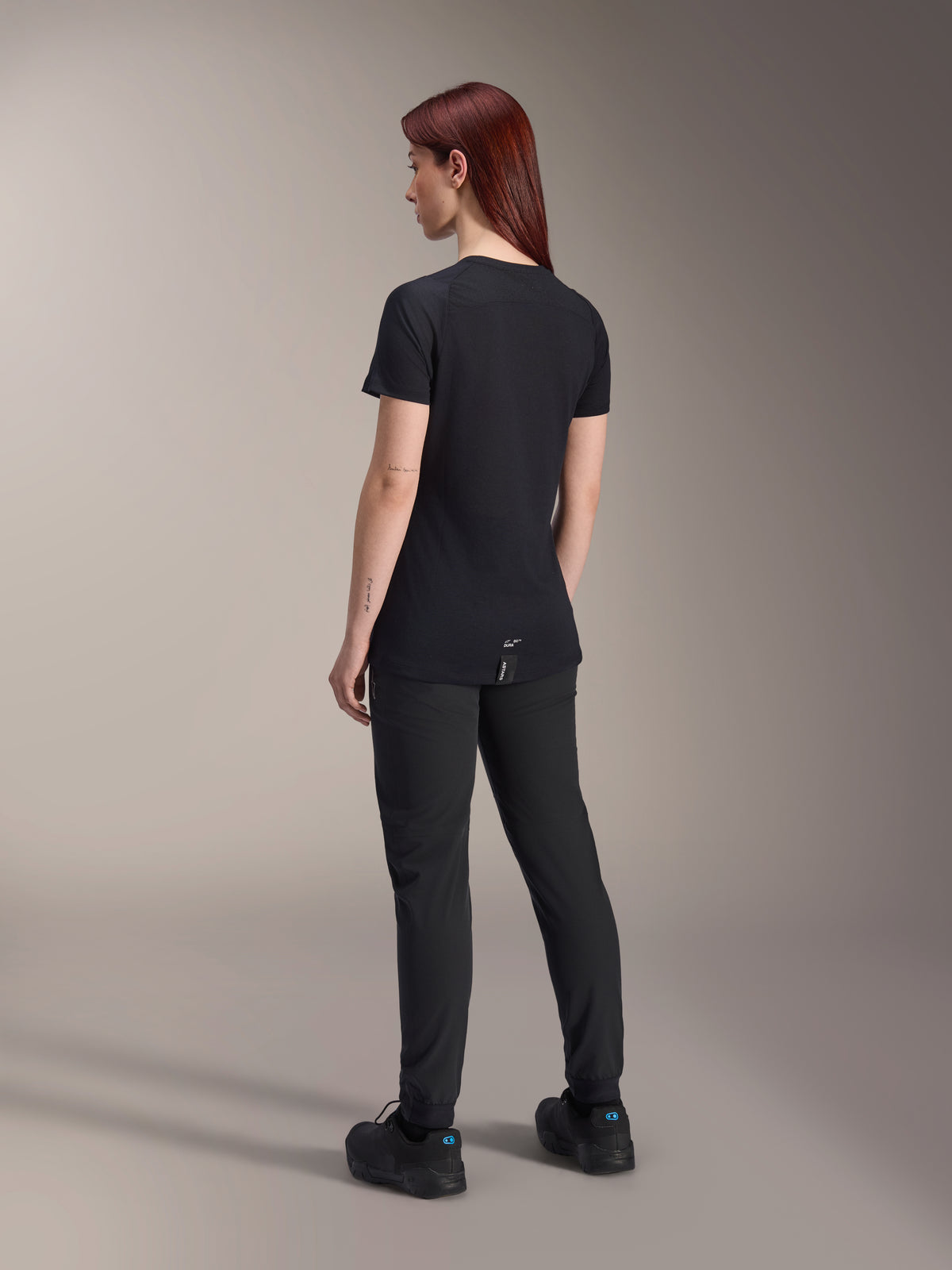 Mannequin femme aux cheveux roux portant le maillot à manches courtes Alpinestars Stella A-Dura Dri Switch en noir, vue de dos, associé à un pantalon et des chaussures de VTT noirs, montrant la coupe technique et le pan arrière allongé pour une couverture optimale à vélo.