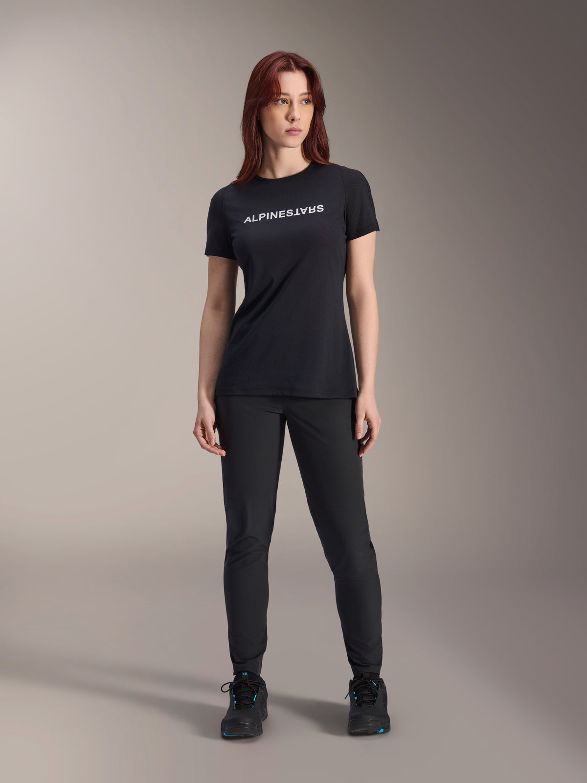 Modèle portant le maillot à manches courtes Alpinestars Stella A-Dura Dri Switch pour femme en noir, avec logo blanc sur la poitrine, associé à un pantalon technique noir et des chaussures de sport noires, photo de studio en pied