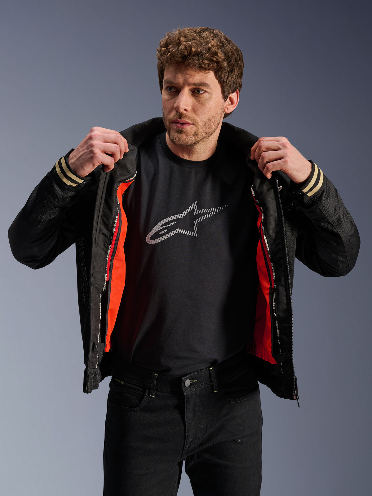 Homme portant la veste Alpinestars Ignite Air noire, dotée d'un corps en maille et d'une doublure coupe-vent amovible orange vif, associée à un t-shirt noir avec logo Astars et un pantalon noir, style moto urbain contemporain