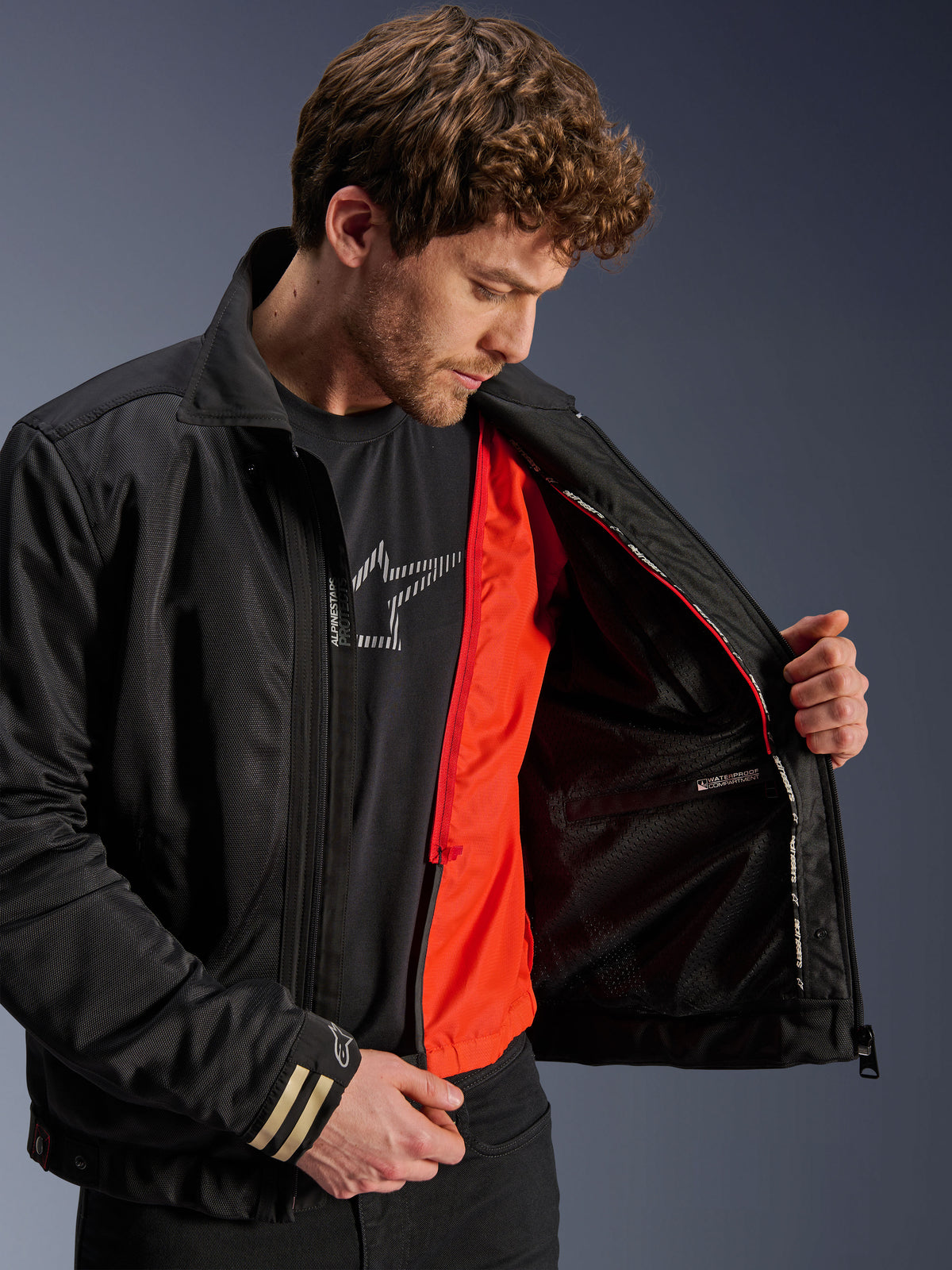 Veste Alpinestars Ignite Air, veste de moto urbaine noire, dotée d'une doublure coupe-vent amovible orange vif et d'un corps en maille, vue intérieure montrant le compartiment imperméable, portée avec un t-shirt noir orné du logo Alpinestars.