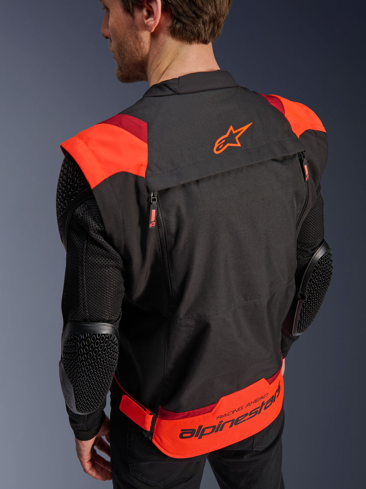 Veste Alpinestars T-Stunt Air, noir, rouge fluo et rouge foncé, vue arrière sur un mannequin, montrant les manches en mesh avec protections de coudes texturées en nid d'abeille, logo étoile Alpinestars sur le haut du dos et marquage « RACING AHEAD » en bas du dos, design modulaire à coupe sportive pour la conduite moto.