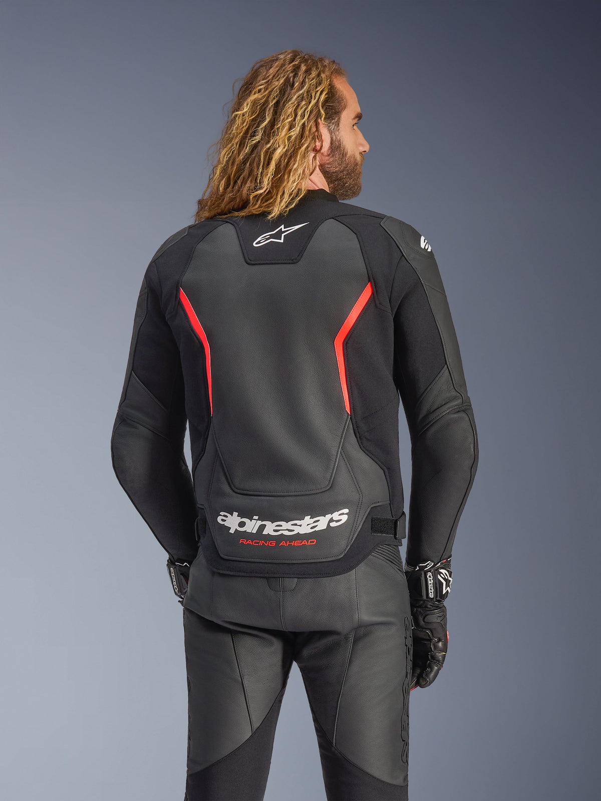 Blouson en cuir Alpinestars GP Force V2, noir et rouge fluo, vue de dos sur un modèle, avec logo Alpinestars blanc et texte 'RACING AHEAD', associé à un pantalon et des gants de moto en cuir noir.