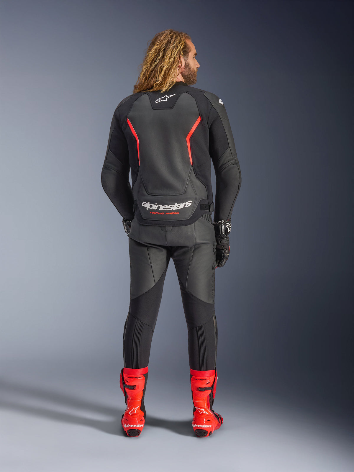 Blouson en cuir Alpinestars GP Force V2, noir et rouge fluo, vu de dos, porté avec un pantalon en cuir noir assorti et des bottes de course rouge vif, avec des lignes d'accentuation rouges et le logo Alpinestars blanc imposant dans le dos, tenue de moto sportive professionnelle.
