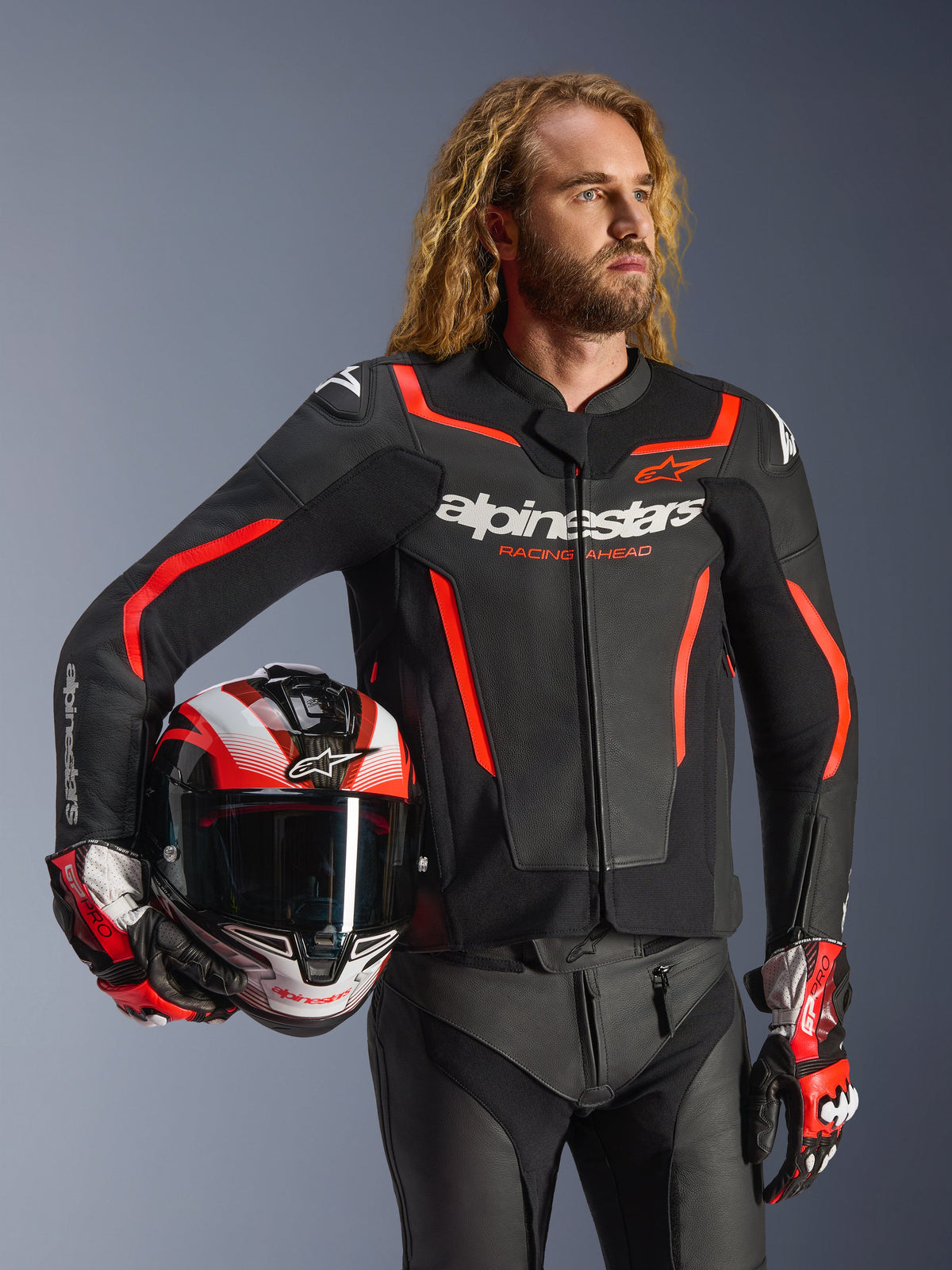 Veste en cuir Alpinestars GP Force V2, noir et rouge fluo, portée par un mannequin homme tenant un casque assorti, avec logos Alpinestars blancs et accents rouges, associée à un pantalon en cuir et des gants de course, équipement de moto sportif pour route et piste.