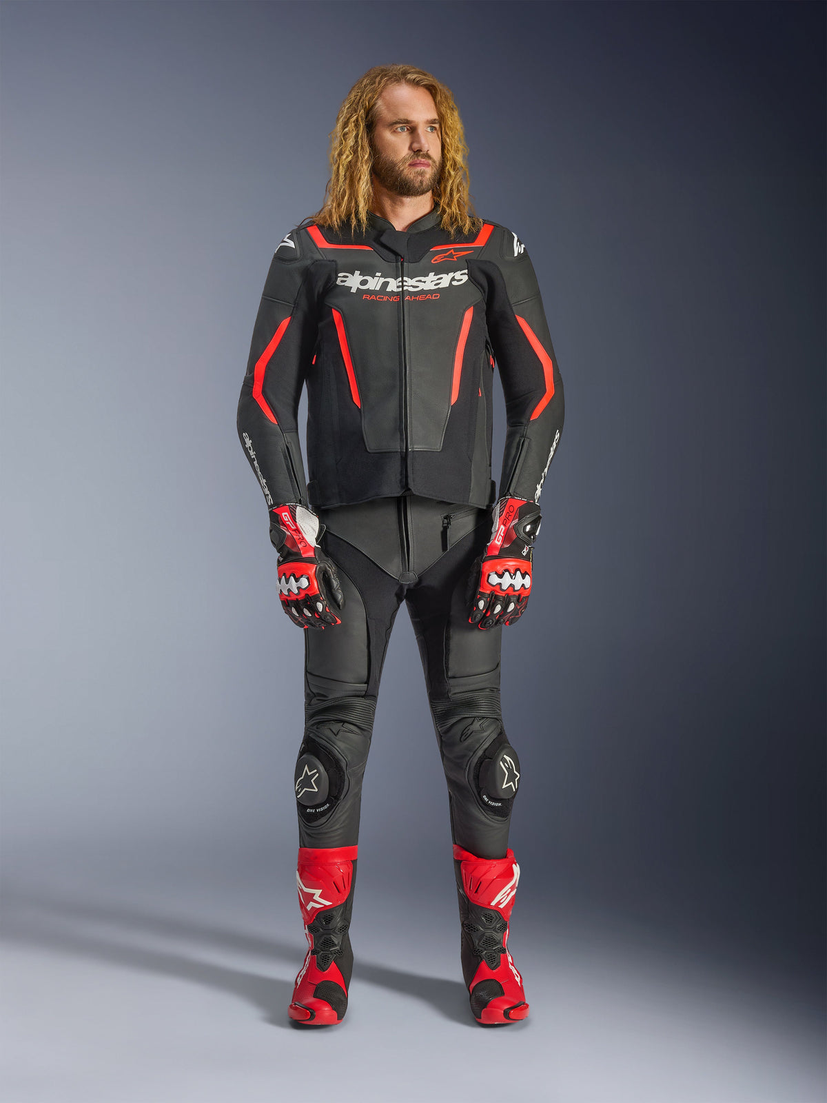 Modèle masculin portant la veste en cuir Alpinestars GP Force V2 en noir et rouge fluo, avec pantalon en cuir assorti, gants de course rouges et bottes de performance rouges, équipement de sport moto professionnel