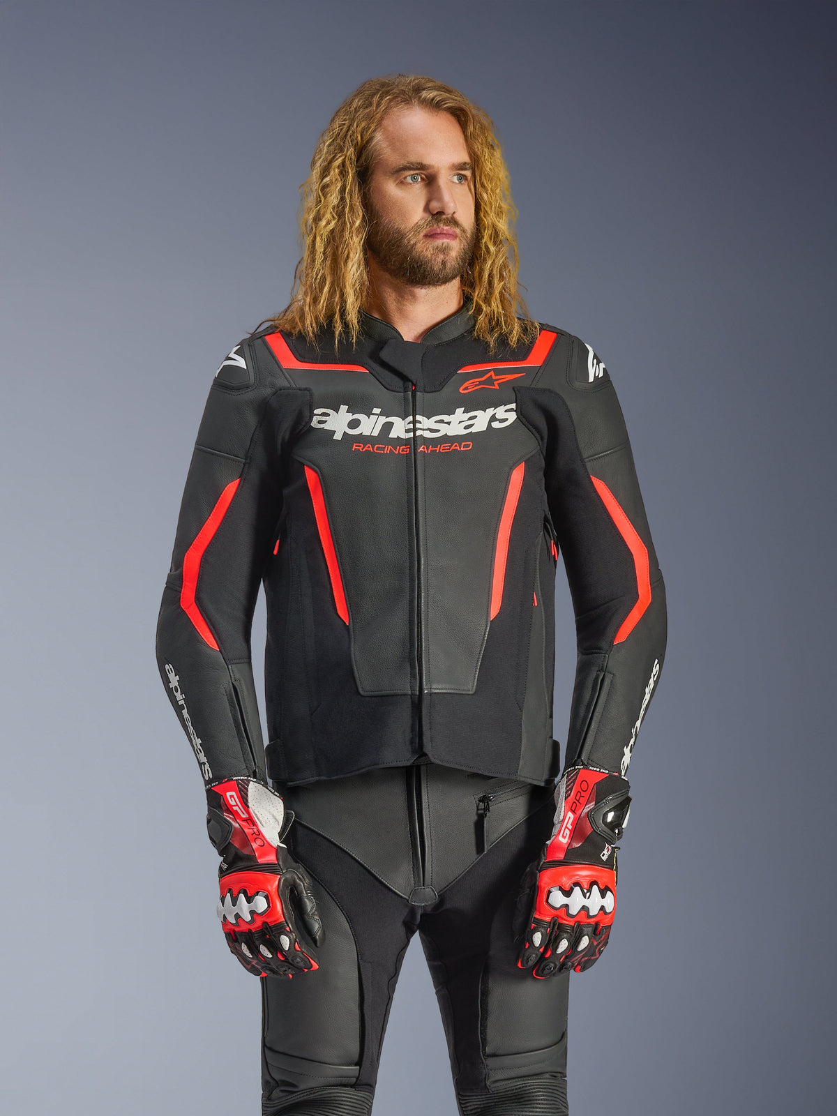 Blouson en cuir Alpinestars GP Force V2, blouson de moto sport, noir et rouge fluo, porté par un mannequin homme aux longs cheveux ondulés, arborant le logo Alpinestars blanc et le texte « RACING AHEAD », avec pantalon et gants en cuir assortis, équipement de pilotage professionnel haute performance