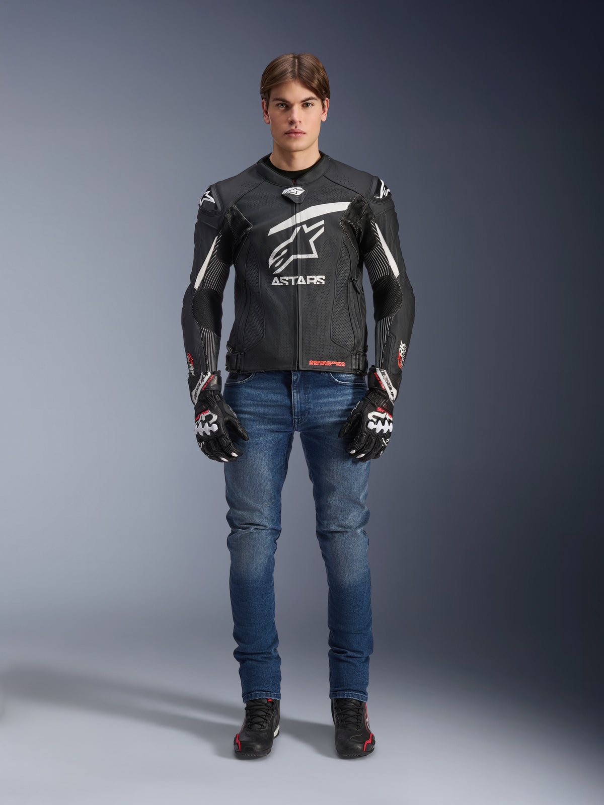 Mannequin homme portant la veste en cuir Alpinestars GP Plus R V4 Rideknit en noir et blanc, associée à un jean de moto bleu, des gants de moto noirs et des chaussures de moto noires, avec un grand logo Astars blanc sur la poitrine, des panneaux en cuir perforé et des bandes de course blanches sur les manches, vue en pied debout