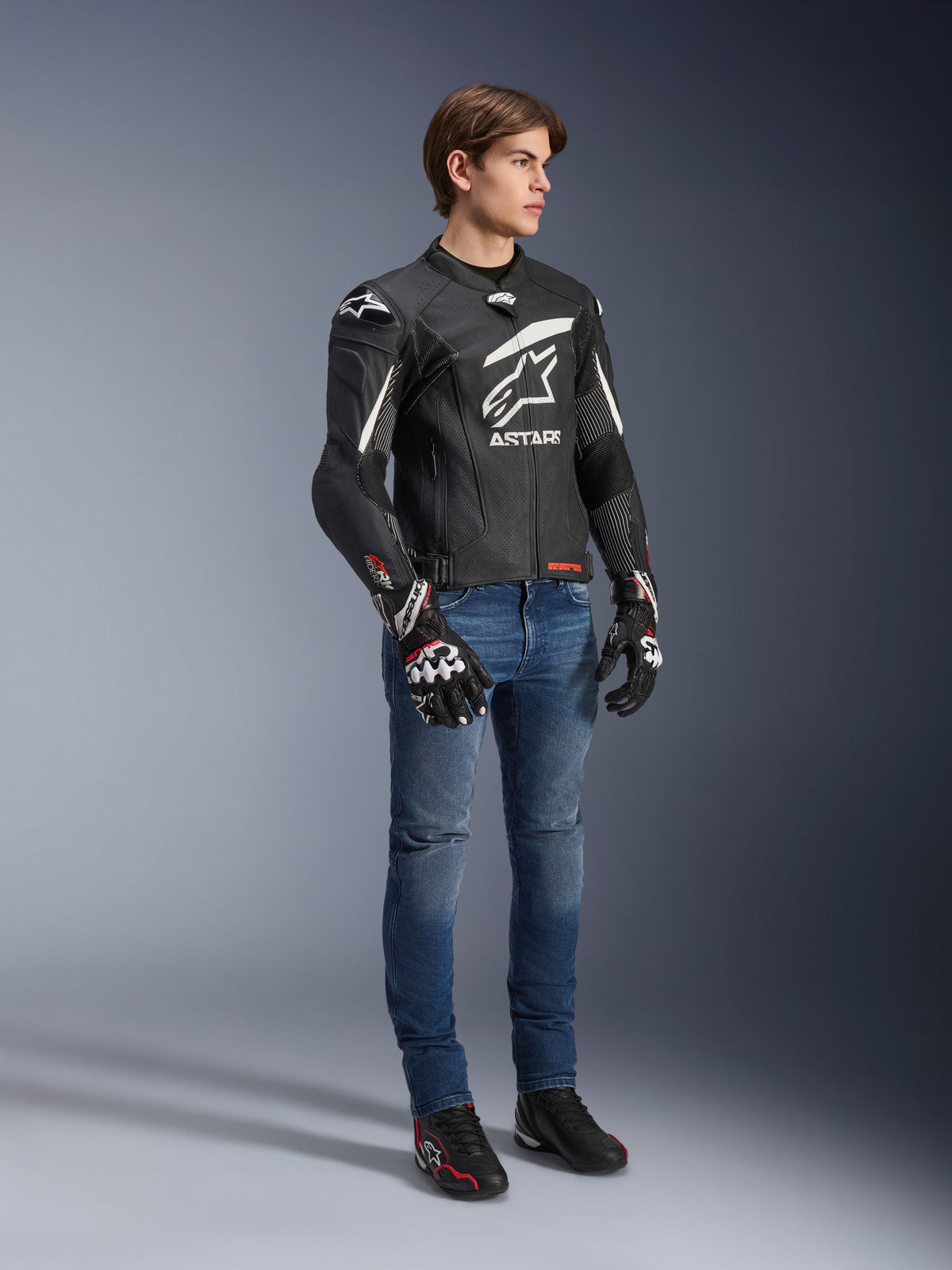 Veste en cuir Alpinestars GP Plus R V4 Rideknit, noir et blanc, veste de moto en cuir portée par un modèle avec un jean bleu et des gants noirs, arborant un logo Astars blanc imposant sur la poitrine, des panneaux perforés et des détails blancs sur les épaules.