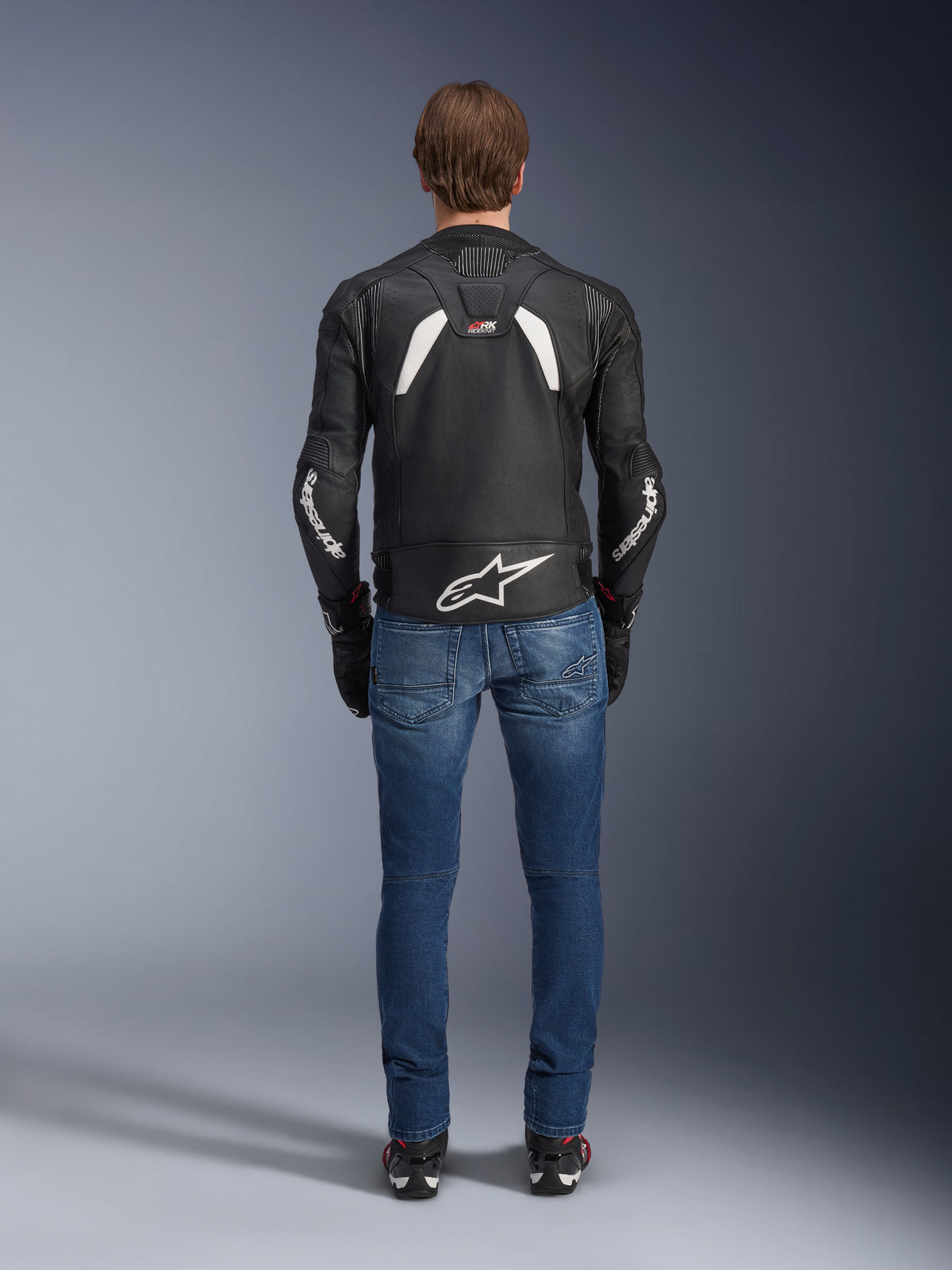 Vue arrière de la veste en cuir Alpinestars GP Plus R V4 Rideknit, noir et blanc, avec un grand logo blanc au bas du dos et un marquage sur les manches, portée avec un jean de moto bleu et des bottes noires, soulignant la construction technique et la coupe sport.