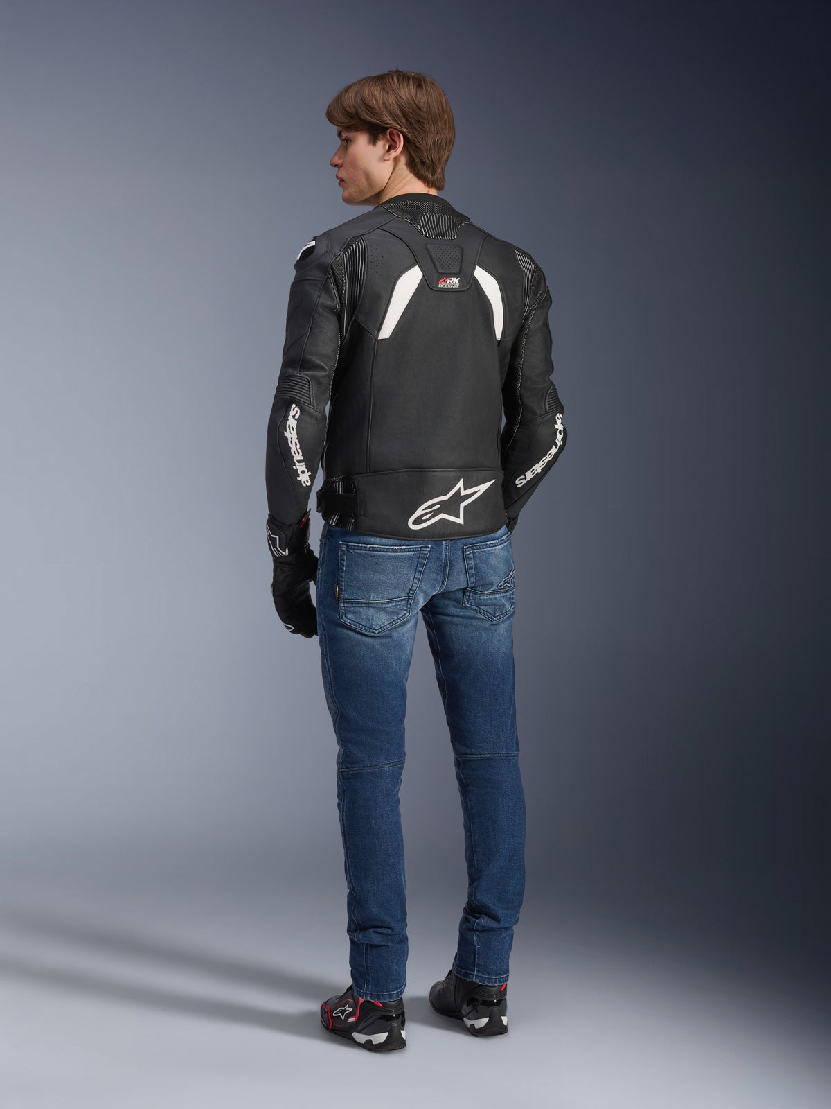 Veste en cuir Alpinestars GP Plus R V4 Rideknit en noir et blanc, vue arrière sur un mannequin, avec marquage Alpinestars blanc sur les manches et un grand logo étoile dans le bas du dos, portée avec un jean bleu et des chaussures de moto noires, équipement technique de moto