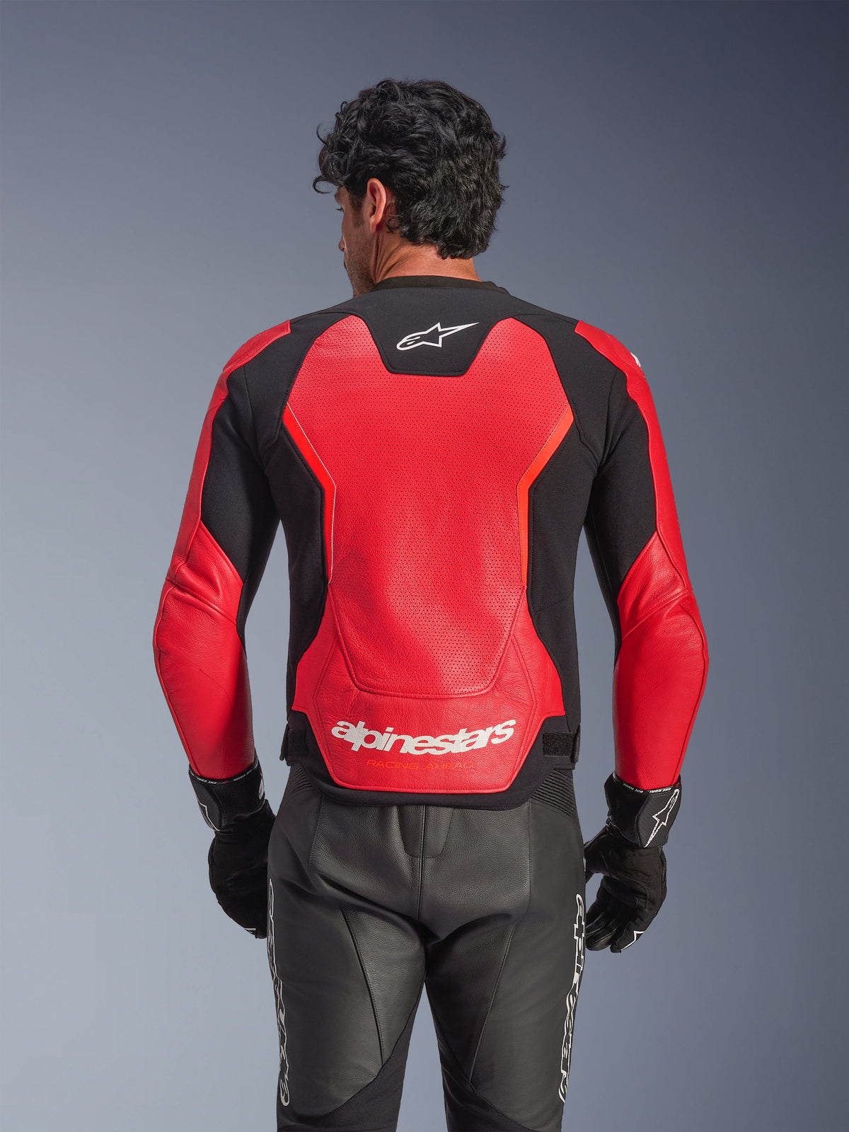 Blouson en cuir Alpinestars GP Force V2 Air en rouge et noir, vue arrière montrant le châssis en cuir perforé pour la circulation de l'air, le logo Alpinestars blanc sur le bas du dos et les panneaux extensibles intégrés, porté avec un pantalon de moto en cuir noir et des gants.