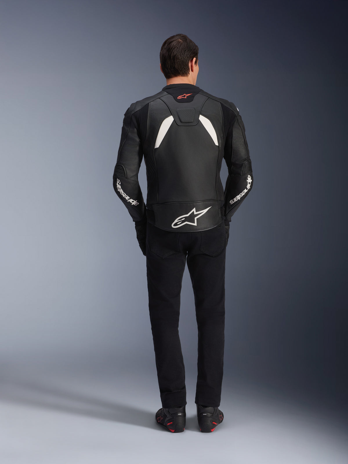 Model viewed from behind wearing the Alpinestars GP Plus V4 Leather Jacket in Black and White, paired with black pants and motorcycle boots, featuring white panels on the shoulders and Alpinestars logo branding on the sleeves and lower back.