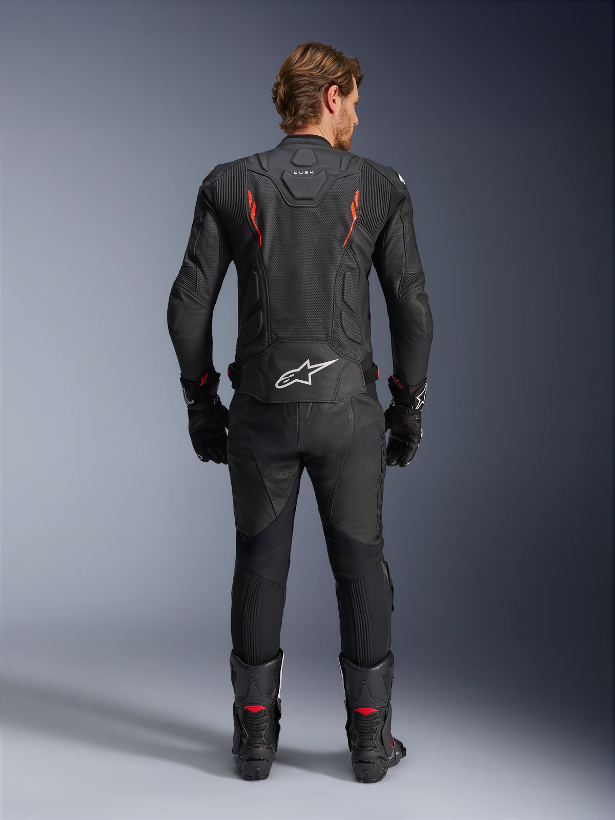 Alpinestars Dusk Airflow Leather Jacket, Black and Red Fluo, rear view showing extensive perforations and large white logo on the lower back, worn with matching leather racing gear including pants, boots, and gloves