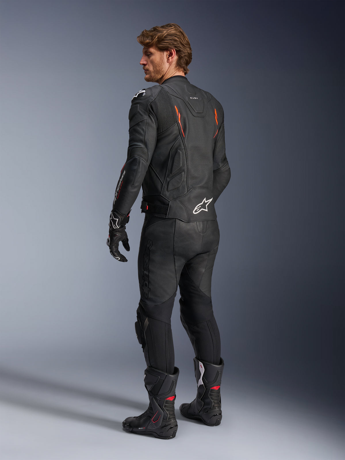 Alpinestars Dusk Airflow Leather Jacket, Black with Red Fluo accents, rear view of a rider in full leather motorcycle gear including matching black pants, gloves, and racing boots, featuring perforated leather and white Alpinestars logos.