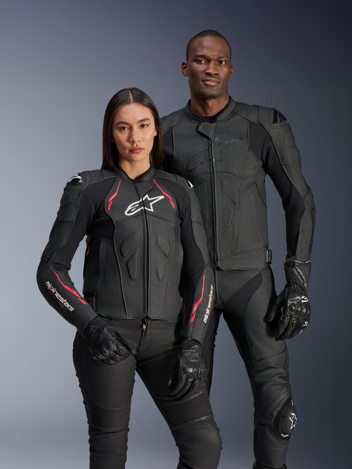 Veste en cuir Alpinestars Stella Dusk, Noir Diva Rose Blanc, portée par un mannequin femme aux côtés d'un mannequin homme en tenue de cuir noir assortie, avec des touches de rose et de blanc, logo étoile blanc sur la poitrine et gants de moto noirs