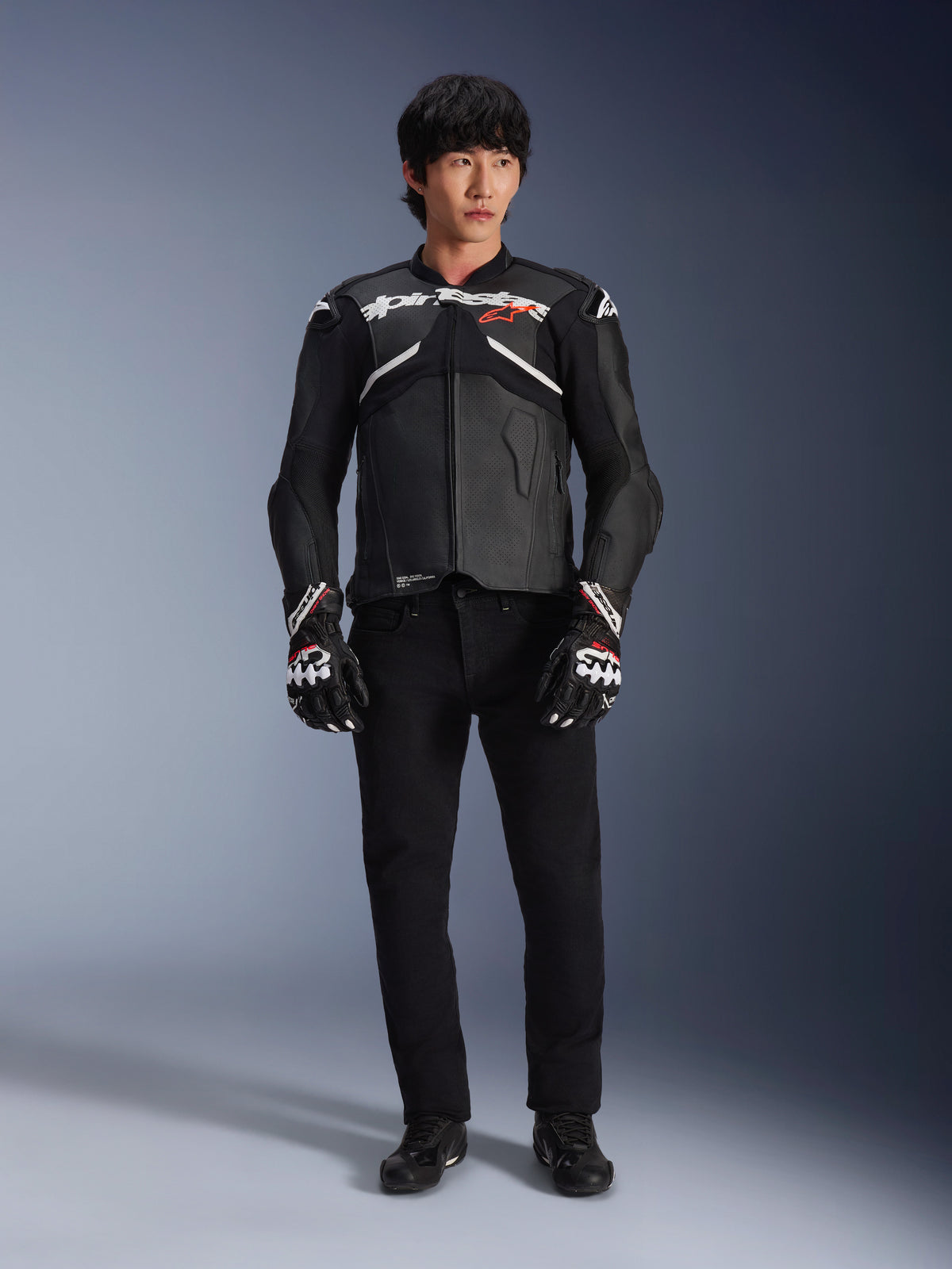 Mannequin portant la veste en cuir Alpinestars Atem V5 en noir et blanc, avec le logo Alpinestars blanc et un logo d'étoile rouge sur la poitrine, associée à un pantalon noir et des gants de course noir et blanc, équipement de moto performant et élégant