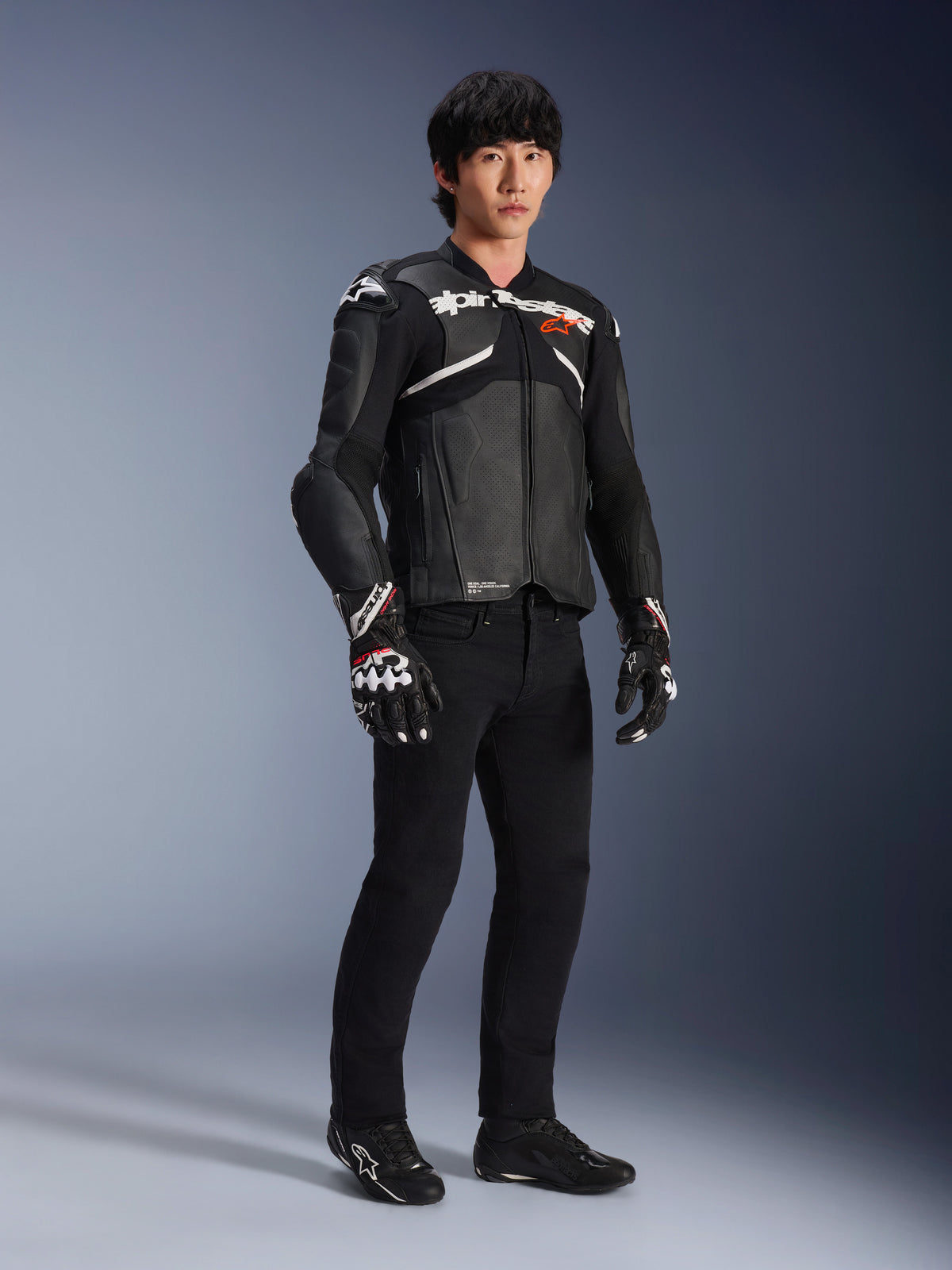 Veste en cuir Atem V5 Alpinestars, veste de course moto, noir et blanc, portée avec un pantalon noir et des gants assortis, avec le logo Alpinestars blanc sur la poitrine et des protections aux épaules.
