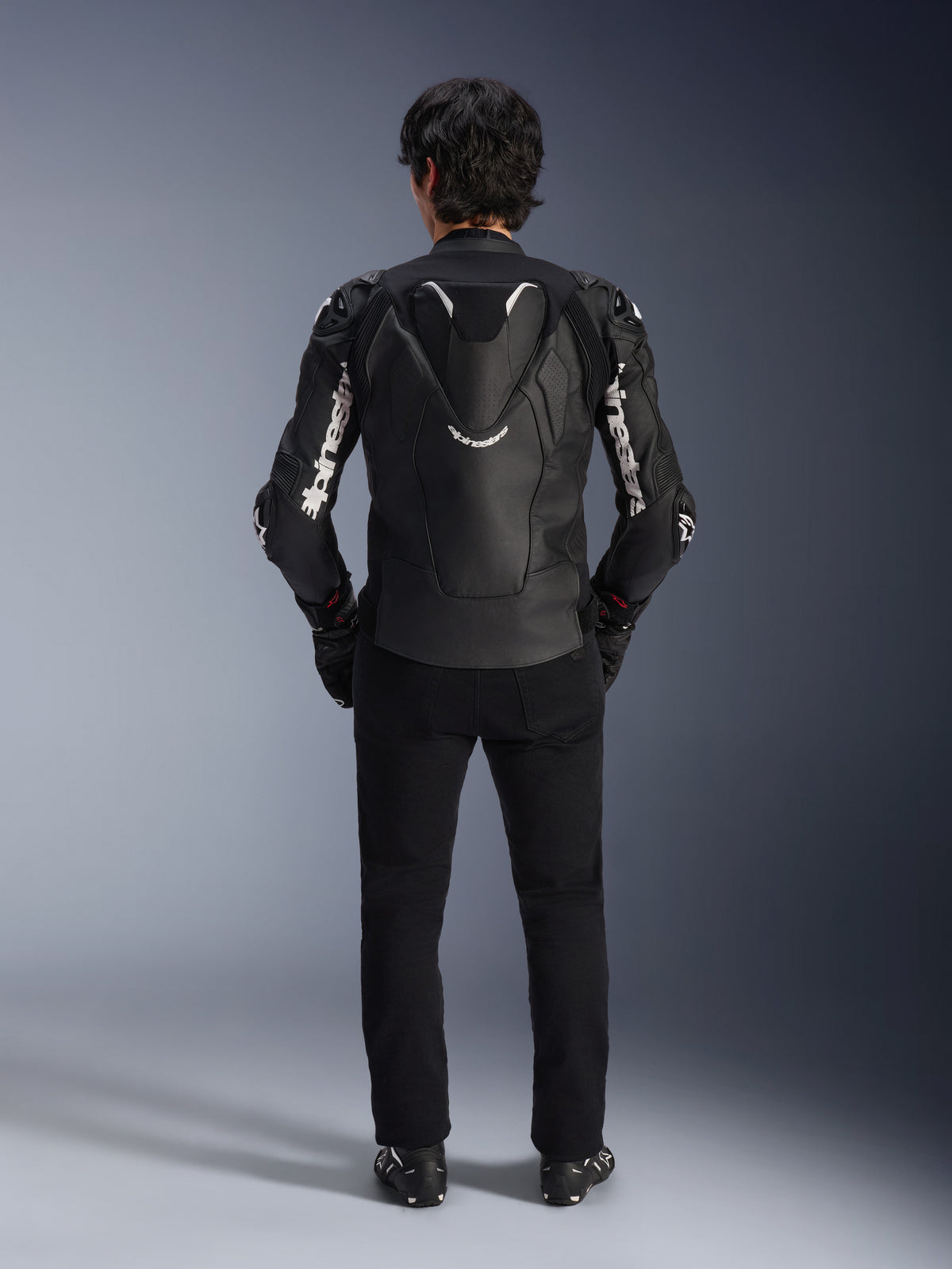 Veste en cuir Alpinestars Atem V5, veste de course moto, noir et blanc, vue arrière sur le pilote, dotée d'une bosse aérodynamique et d'un marquage Alpinestars blanc sur les manches et le dos, construction en cuir technique, portée avec un pantalon noir et des bottes de moto