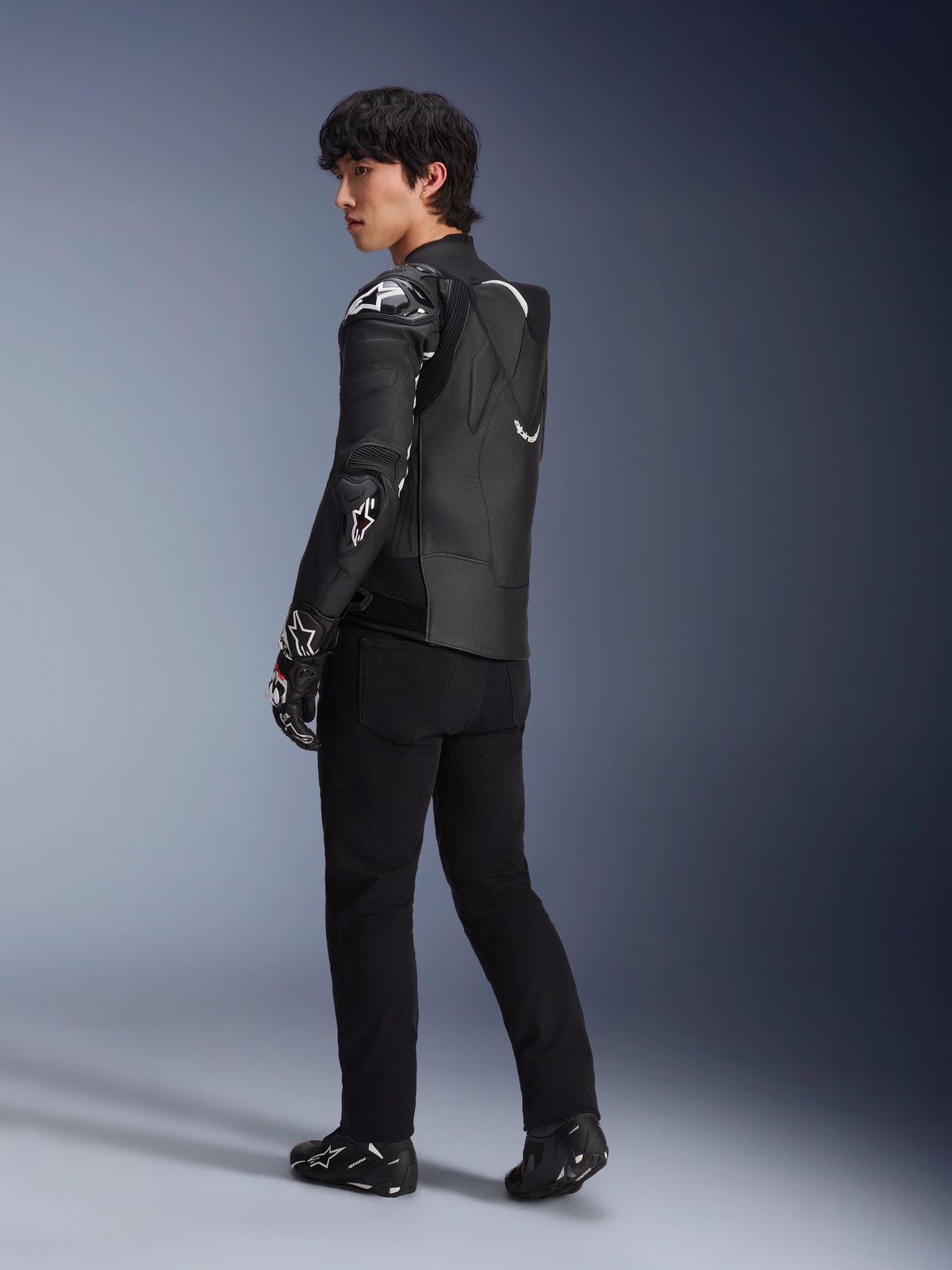 Modèle masculin portant la veste en cuir Alpinestars Atem V5 en noir et blanc, vue de trois-quarts arrière montrant la bosse aérodynamique et les protections DFS RR, assortie d'un pantalon noir, de gants de course et de bottes, équipement de moto de course professionnel