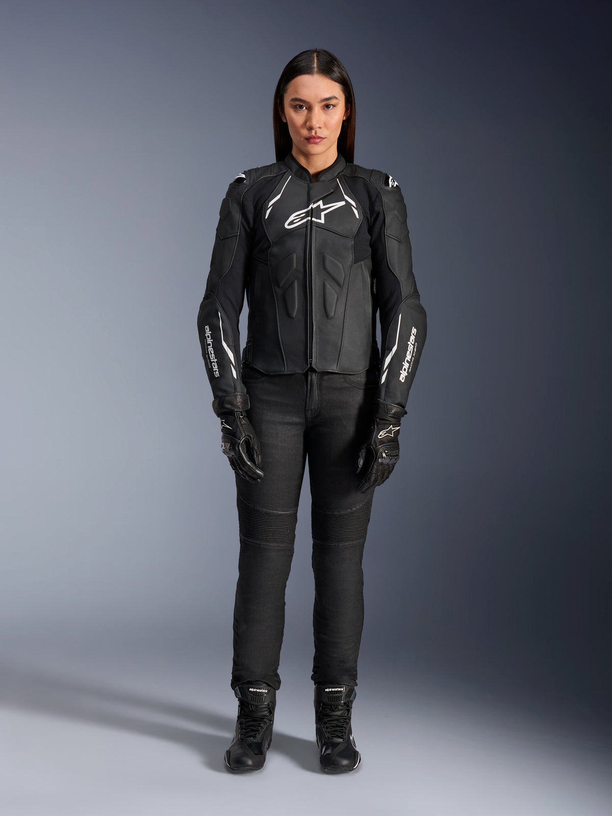 Veste en cuir pour femme Alpinestars Stella Dusk, noir et blanc, portée par un mannequin avec un pantalon et des bottes noirs assortis, vue de face, logos Alpinestars blancs sur la poitrine et les manches, équipement de protection en cuir bovin premium pour femme