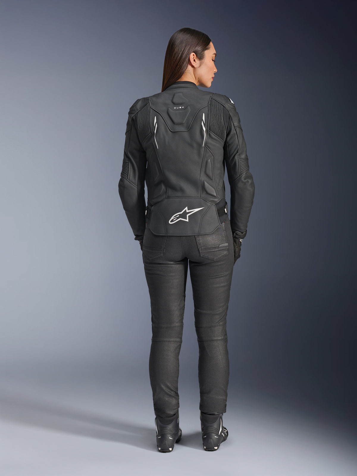 Veste en cuir pour femme Stella Dusk d'Alpinestars, noire, vue arrière avec logo Alpinestars blanc en bas du dos et marquage 'DUSK' en haut du dos, portée avec un pantalon de moto et des bottes noirs, mettant en valeur la coupe féminine ajustée et la construction protectrice en cuir.