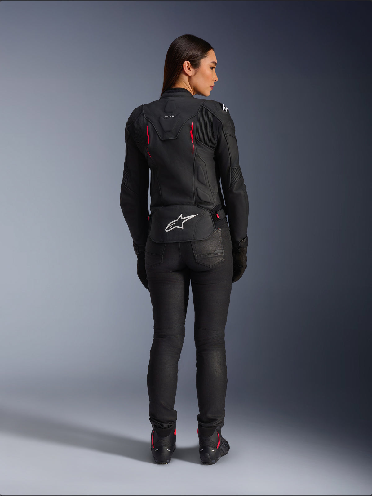 Veste en cuir Alpinestars Stella Dusk, vue de dos, cuir noir avec accents Diva Pink et blancs, arborant le logo Alpinestars blanc en bas du dos et des détails de ventilation roses, portée avec un pantalon de moto et des bottes noirs, équipement de moto sportif haut de gamme pour femmes