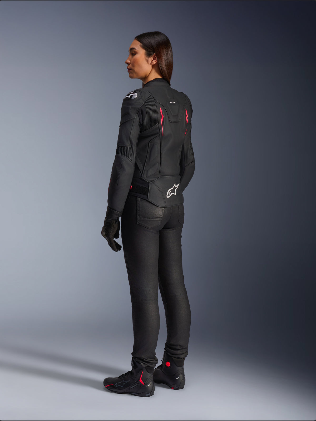 Veste en cuir pour femme Stella Dusk, Noir Diva Rose Blanc, vue arrière de trois-quarts sur une motarde, portée avec un pantalon de moto noir et des bottes noires, dotée d'une construction en cuir élégante avec des accents roses et un logo Alpinestars blanc dans le bas du dos.