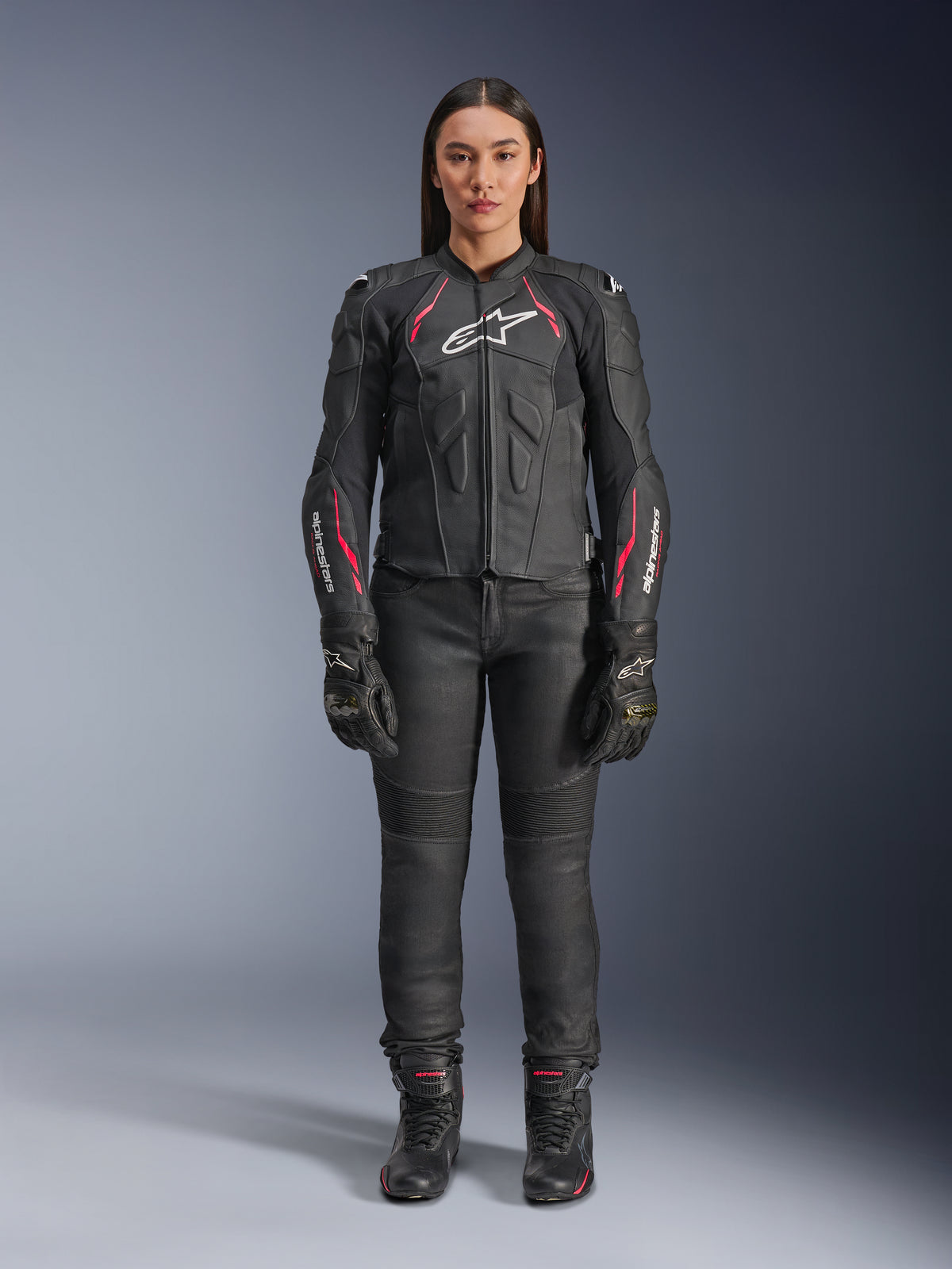 Une femme portant la veste en cuir Alpinestars Stella Dusk en coloris Noir Diva Rose Blanc, avec un logo blanc sur la poitrine et des touches de rose sur les épaules et les manches, accompagnée d'un pantalon de moto, de gants et de bottes noirs.