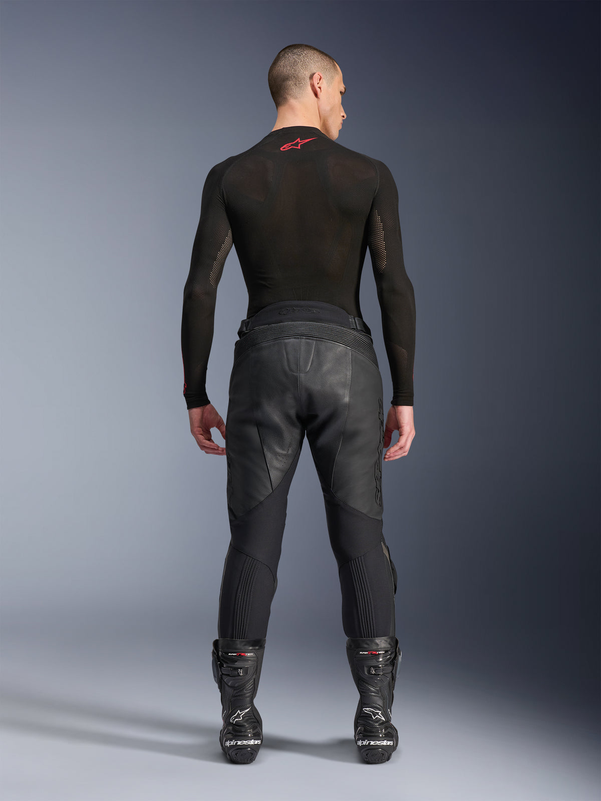 Pantalon en cuir Alpinestars Missile V3, noir, vue arrière sur un modèle portant un haut de compression noir à manches longues et des bottes de course, construction en cuir de première qualité avec taille haute surélevée et zones extensibles, équipement de course moto professionnel