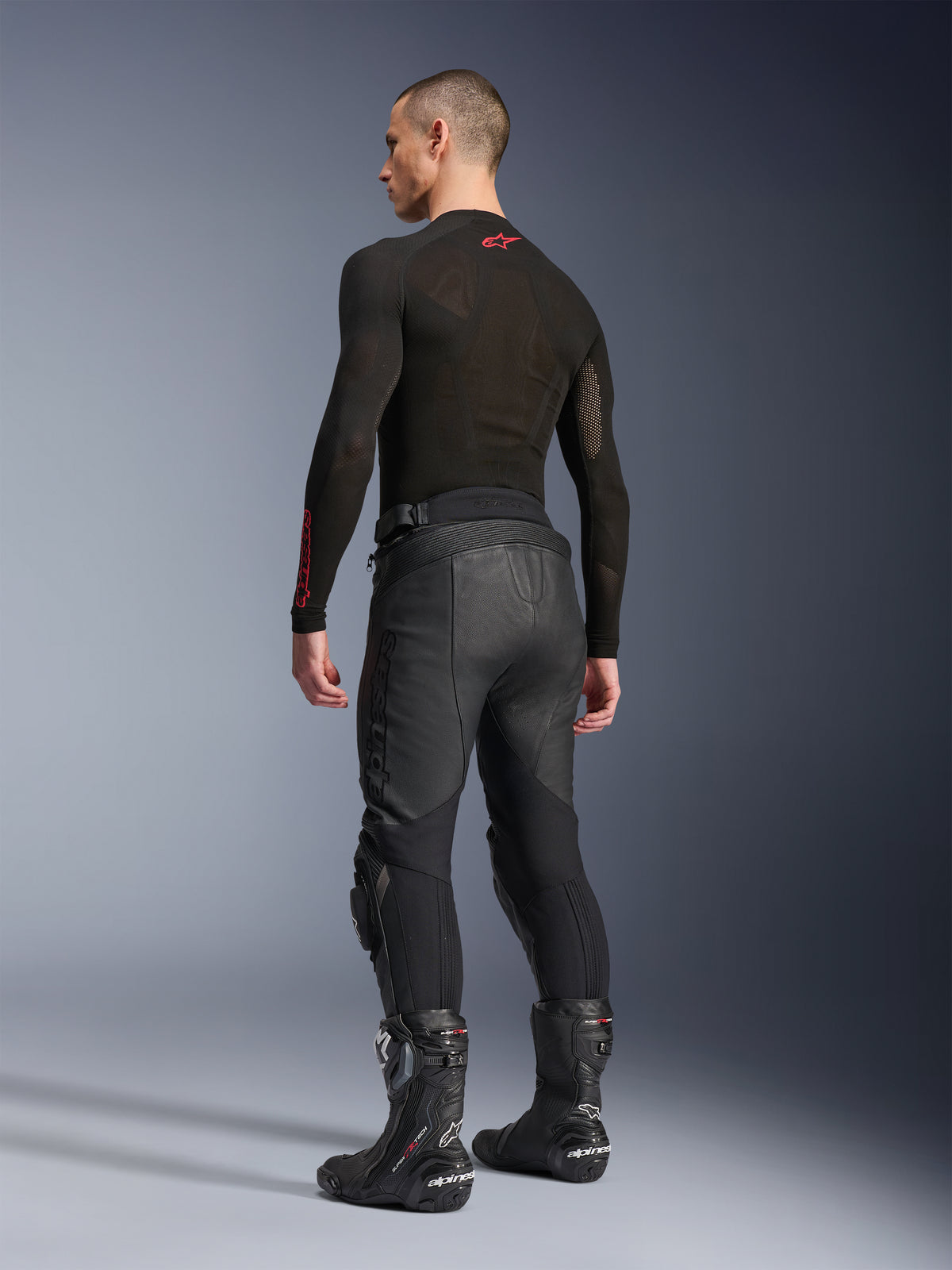 Pantalon en cuir Alpinestars Missile V3, noir, vue arrière sur modèle, porté avec un sous-vêtement technique à manches longues noir et des bottes de course moto noires, doté de panneaux extensibles en cuir accordéon et d'une conception pré-courbée pour un ajustement ergonomique.