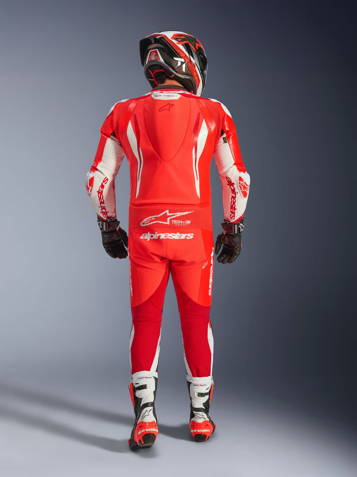 Vue arrière d'un pilote portant la combinaison en cuir une pièce Alpinestars GP Tech V5 en rouge fluo, rouge vif et blanc. Combinaison en cuir haute performance avec bosse aérodynamique, détails blancs et logo Alpinestars, associée à un casque assorti, des gants noirs et des bottes de course blanches et rouges.