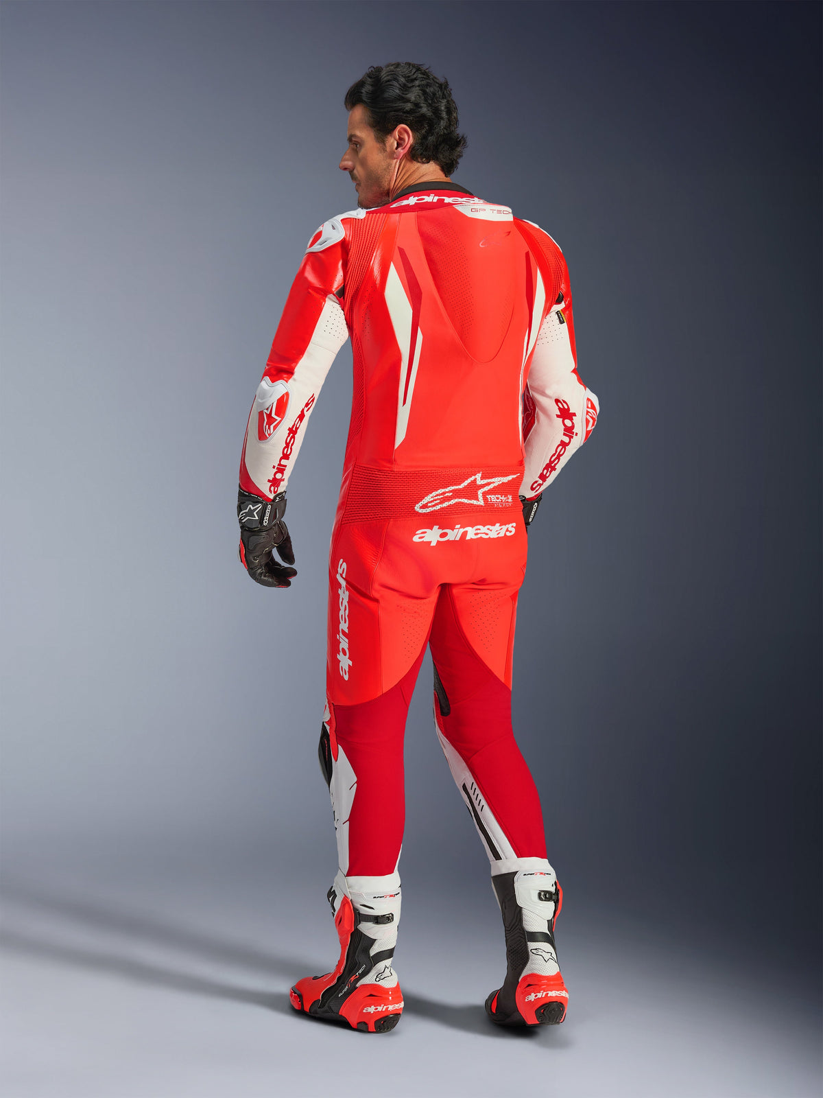 Combinaison en cuir Alpinestars GP Tech V5 1PC, rouge fluo, rouge vif et blanc, vue de dos montrant la bosse aérodynamique et les logos Alpinestars, portée avec des gants de course noirs et des bottes de course blanches et rouges, équipement de course moto professionnel