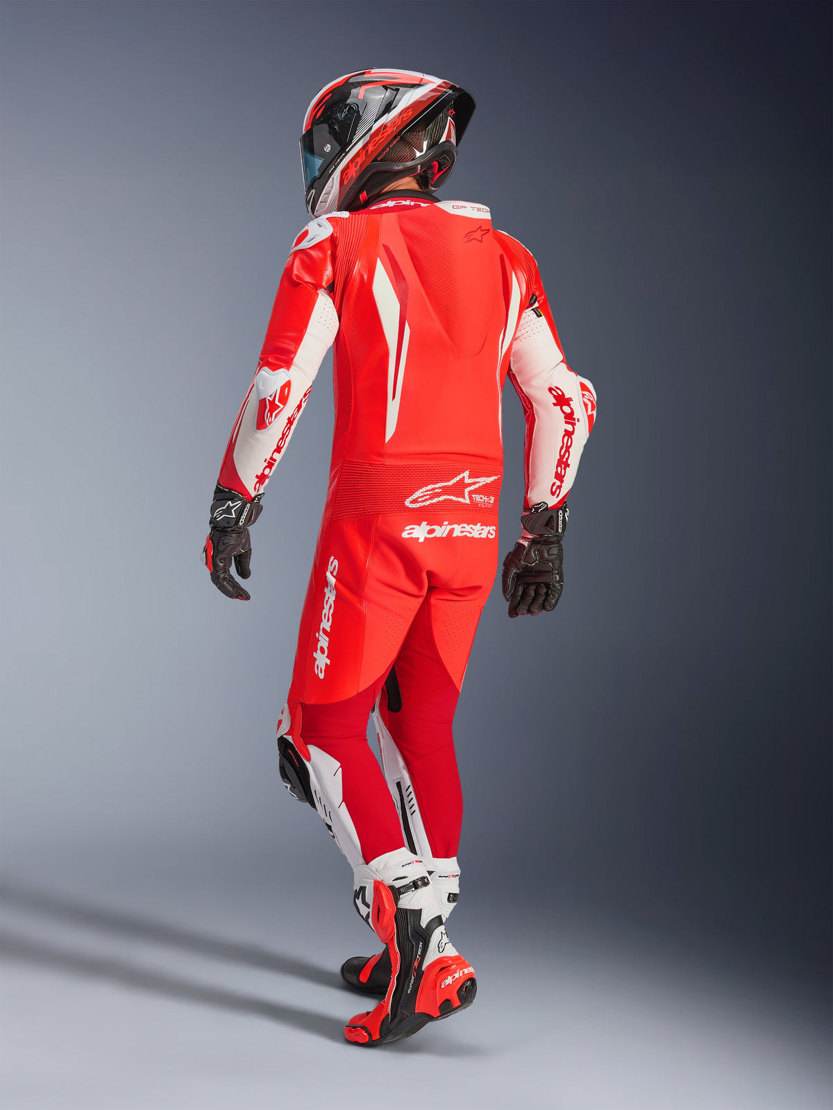 Combinaison en cuir Alpinestars GP Tech V5, Rouge Fluo, Rouge Vif et Blanc, vue arrière d'un motocycliste en tenue de course complète, dotée d'une bosse aérodynamique et de panneaux blancs sur les manches et les jambes, associée à un casque assorti et des bottes de course rouges, conception haute performance pour la piste.