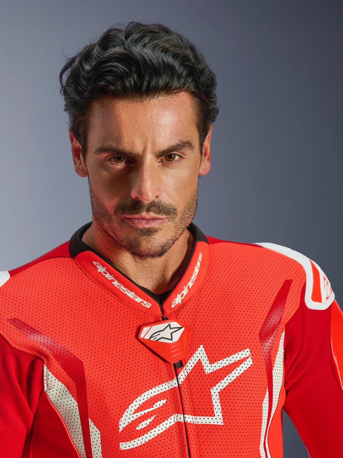 Combinaison en cuir une pièce Alpinestars GP Tech V5, combinaison de course moto, rouge fluo, rouge vif et blanc, portrait en gros plan d'un pilote, poitrine en cuir perforé avec logo Alpinestars blanc, col de course haute performance et design aérodynamique.