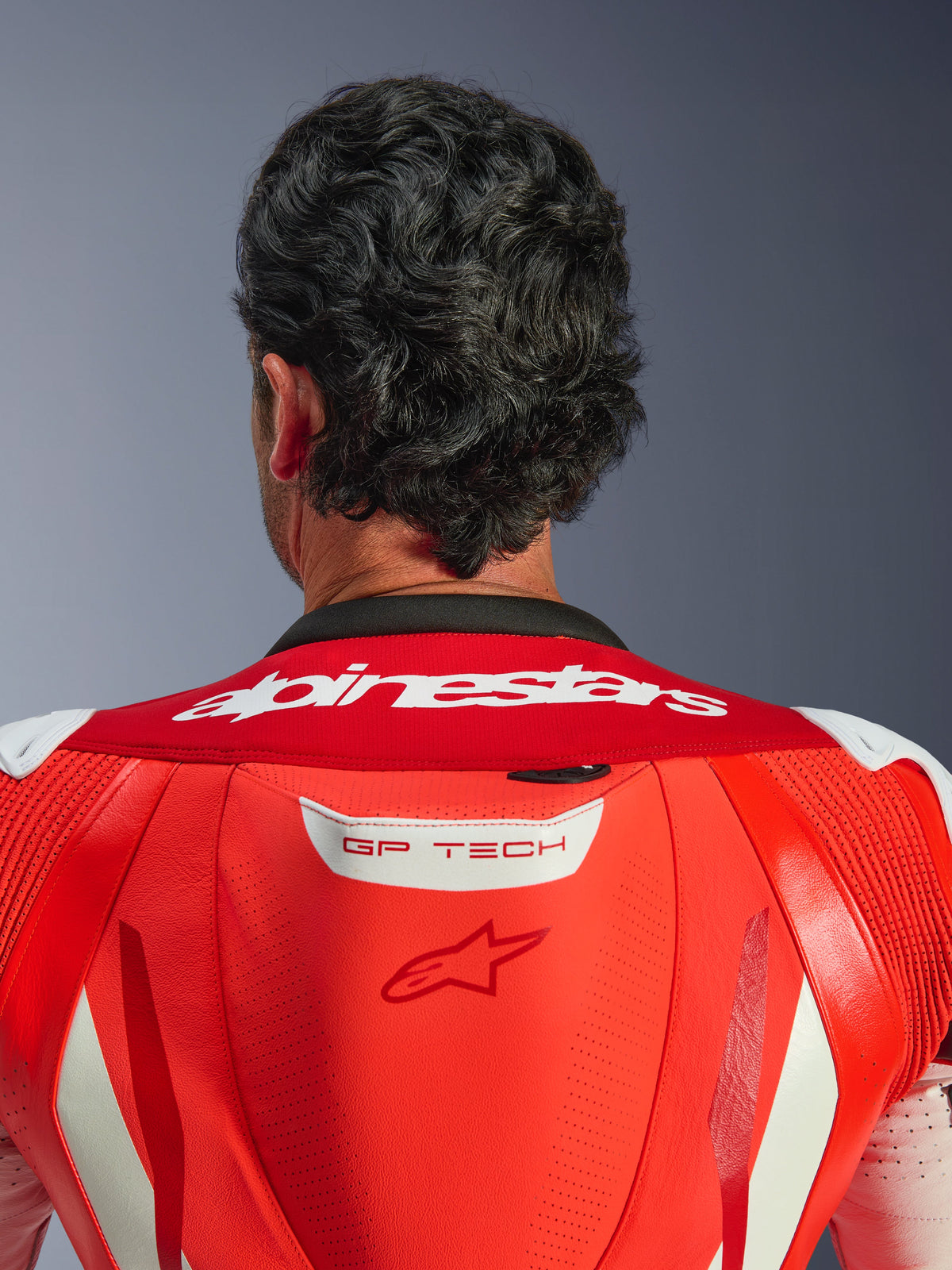 Gros plan sur le haut du dos et la nuque de la combinaison en cuir une pièce GP Tech V5, Rouge Fluo et Rouge Vif, présentant le lettrage Alpinestars blanc sur le col et le logo GP TECH sur la bosse aérodynamique, portée par un pilote.